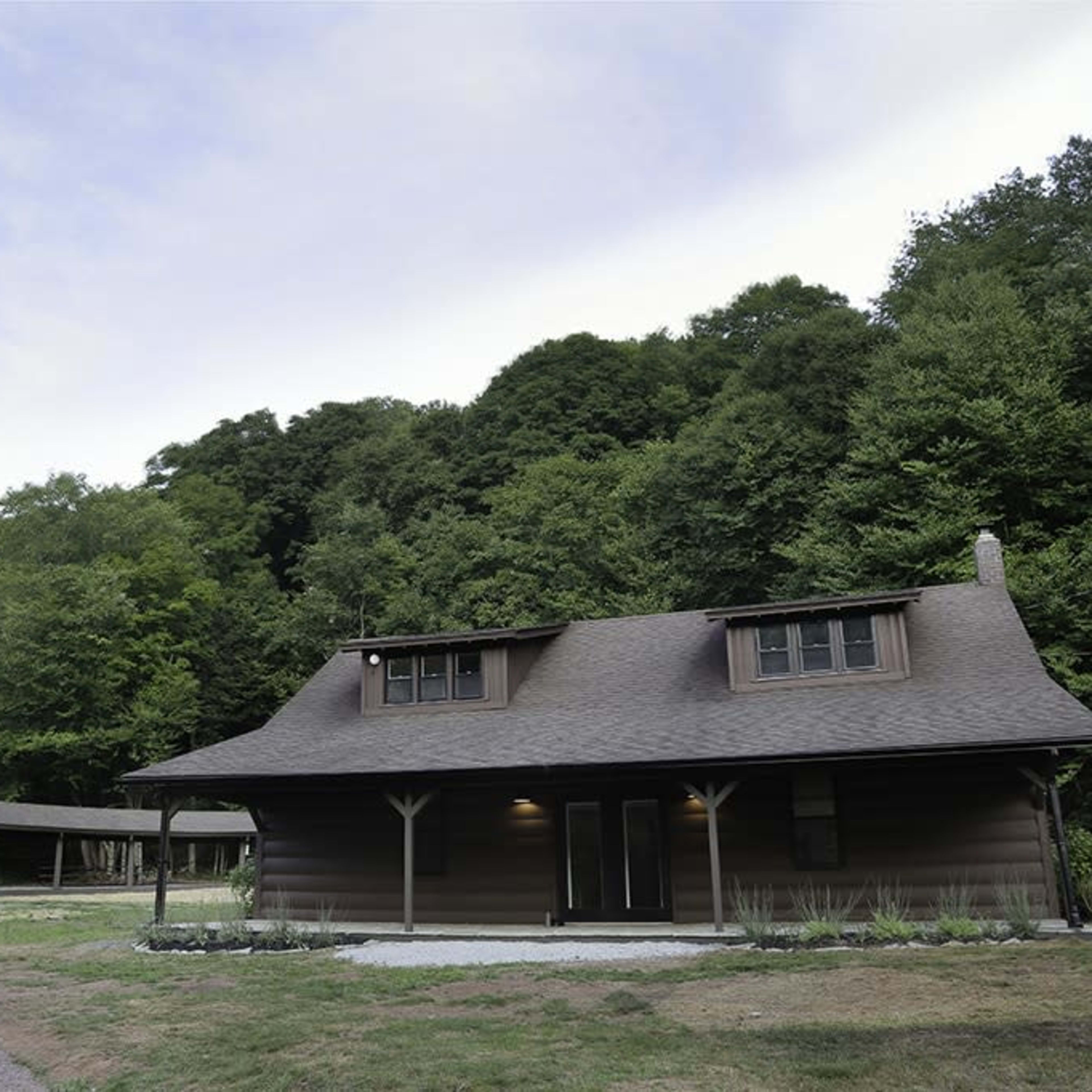 A cabin-style house is situated on a gravel driveway, surrounded by greenery and trees in the background.