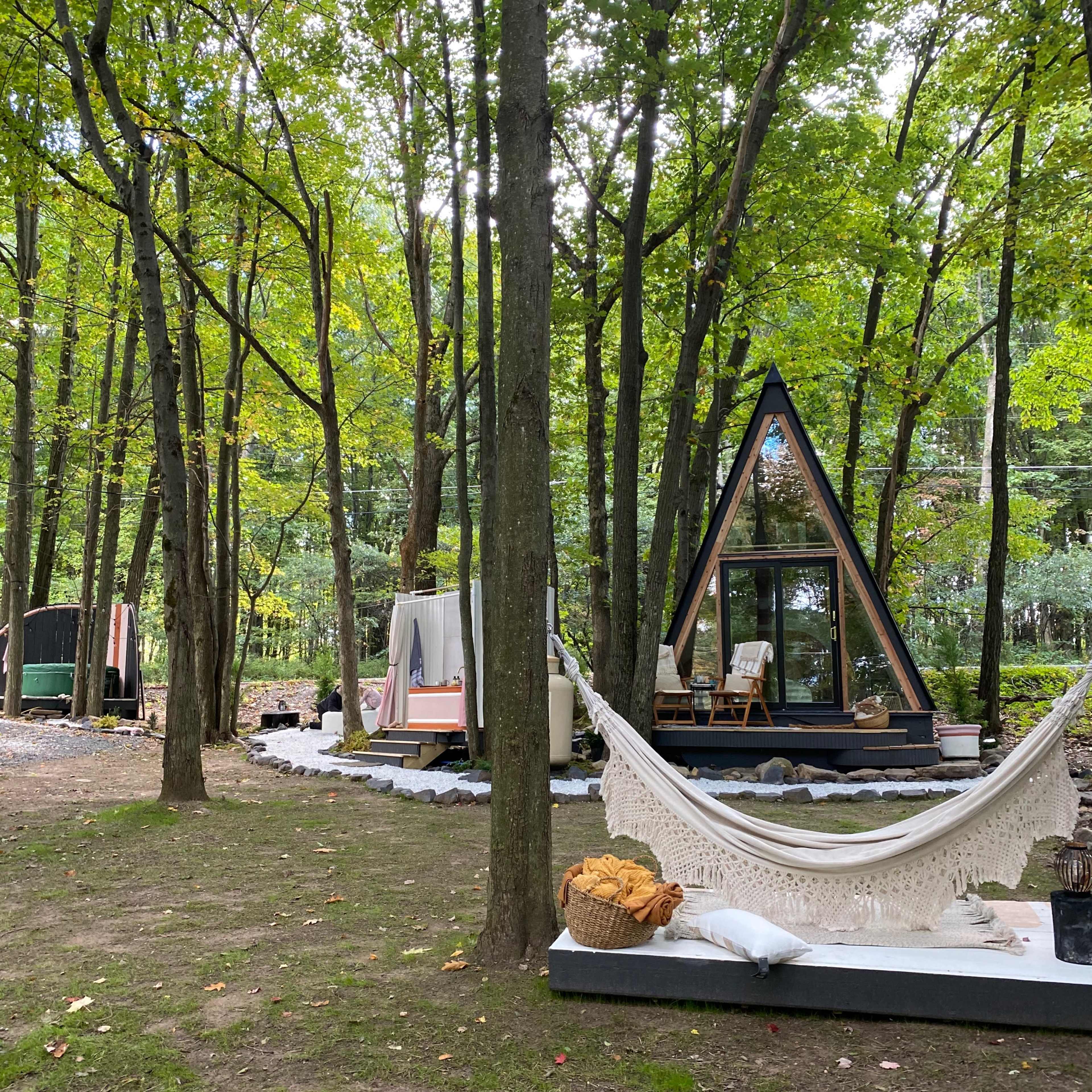 A modern A-frame cabin with large windows is nestled among trees in a wooded area, featuring a hammock and outdoor seating.