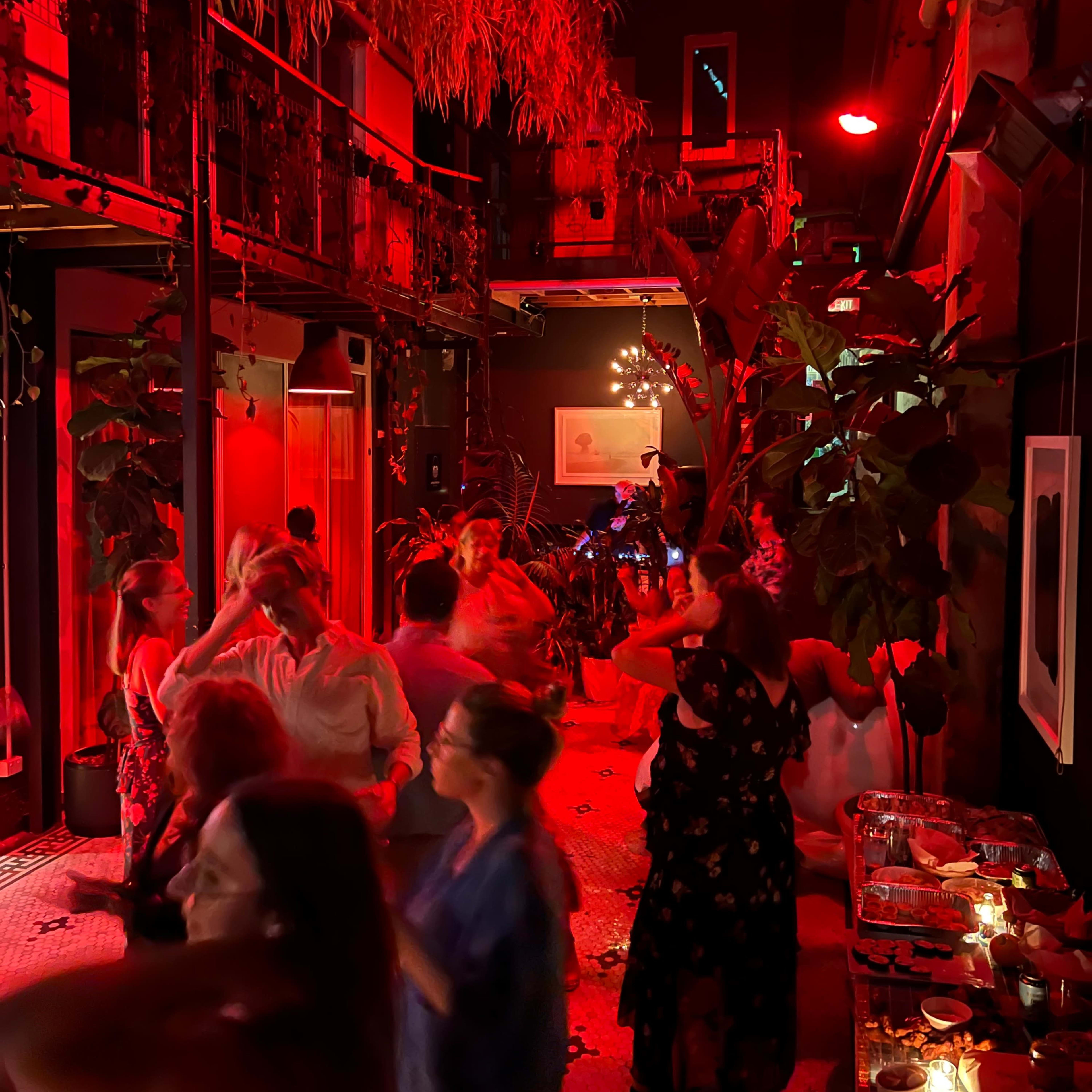 A lively party scene with people dancing under red lighting in a decorated indoor space filled with plants.