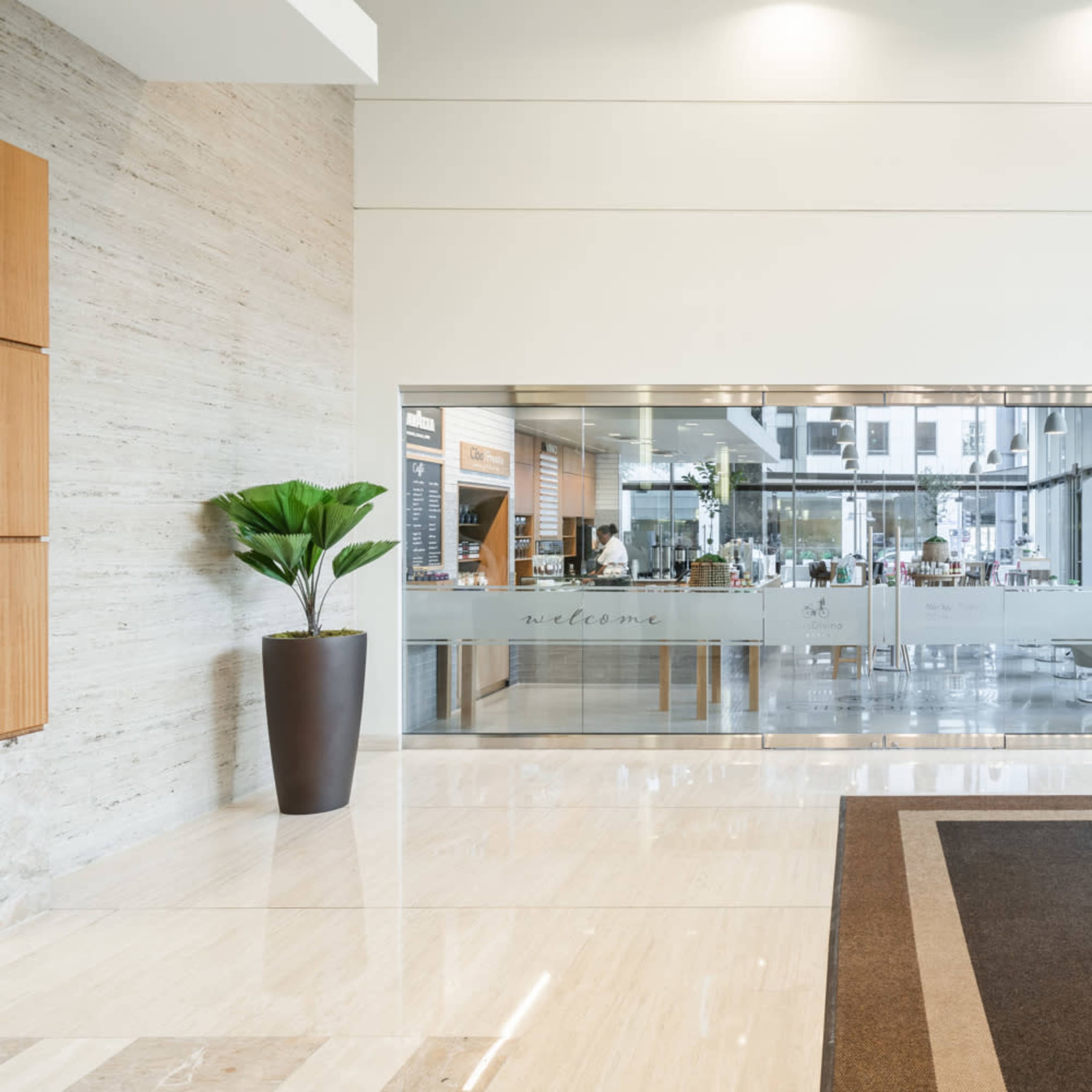 A modern interior lobby with a large glass facade revealing a dining area, beige marble walls, and a potted plant near the entrance.