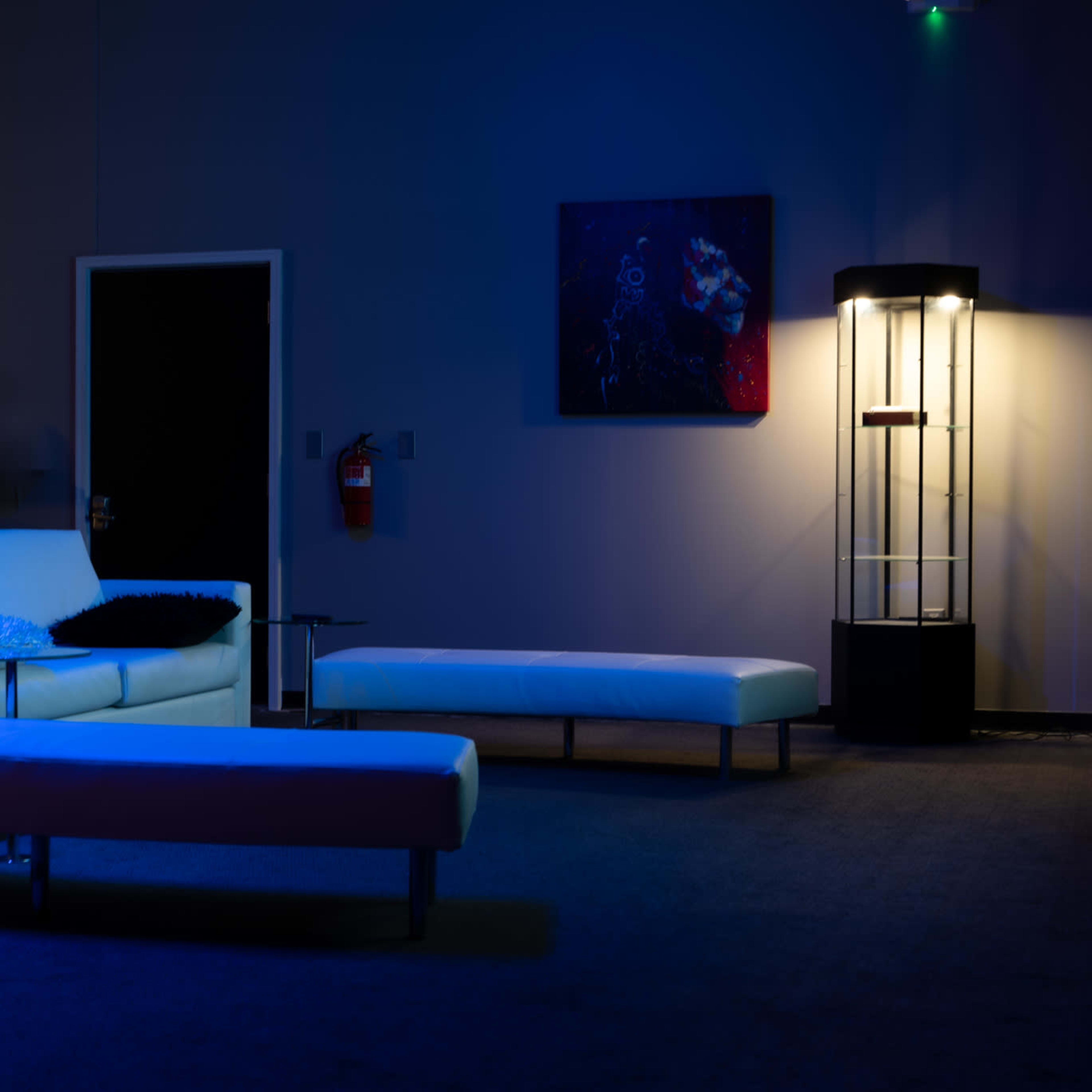 A minimalist room with a white couch and benches under blue lighting, accompanied by a wall-mounted artwork and a standing lamp.