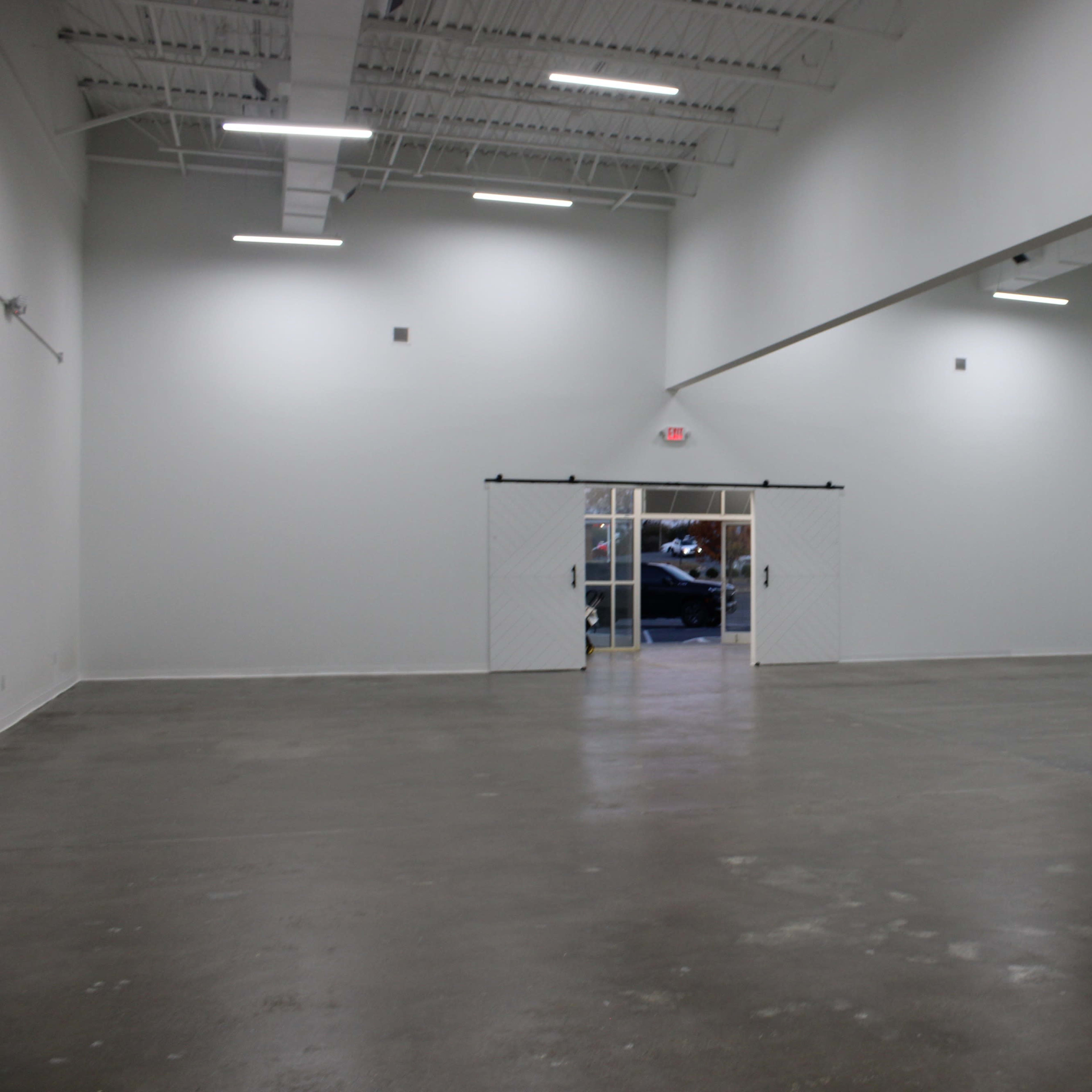 A large, empty commercial space with white walls and a concrete floor, featuring double doors at the far end.