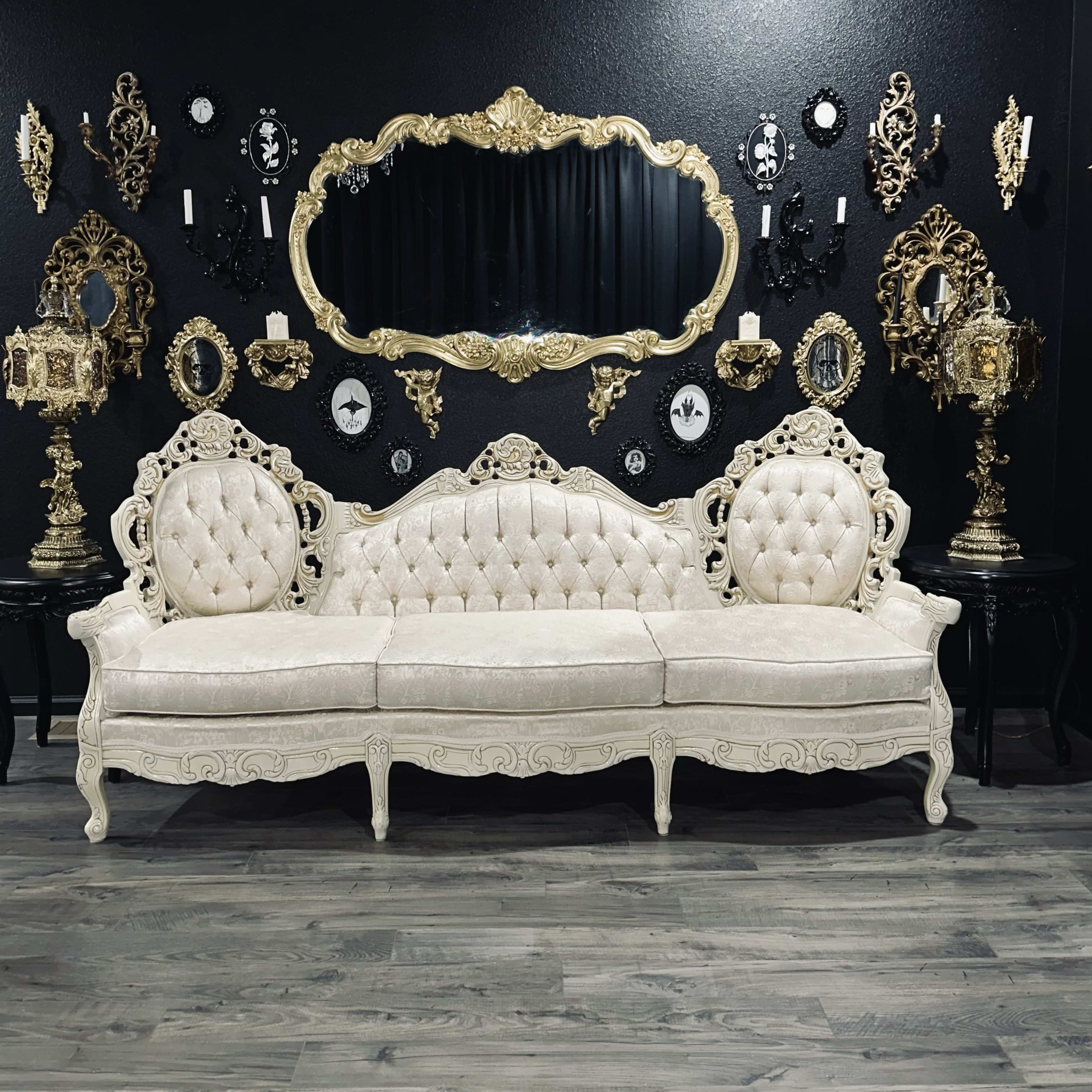 A vintage ornate white sofa is positioned in front of a large decorative mirror, surrounded by framed artwork and antique wall decorations.