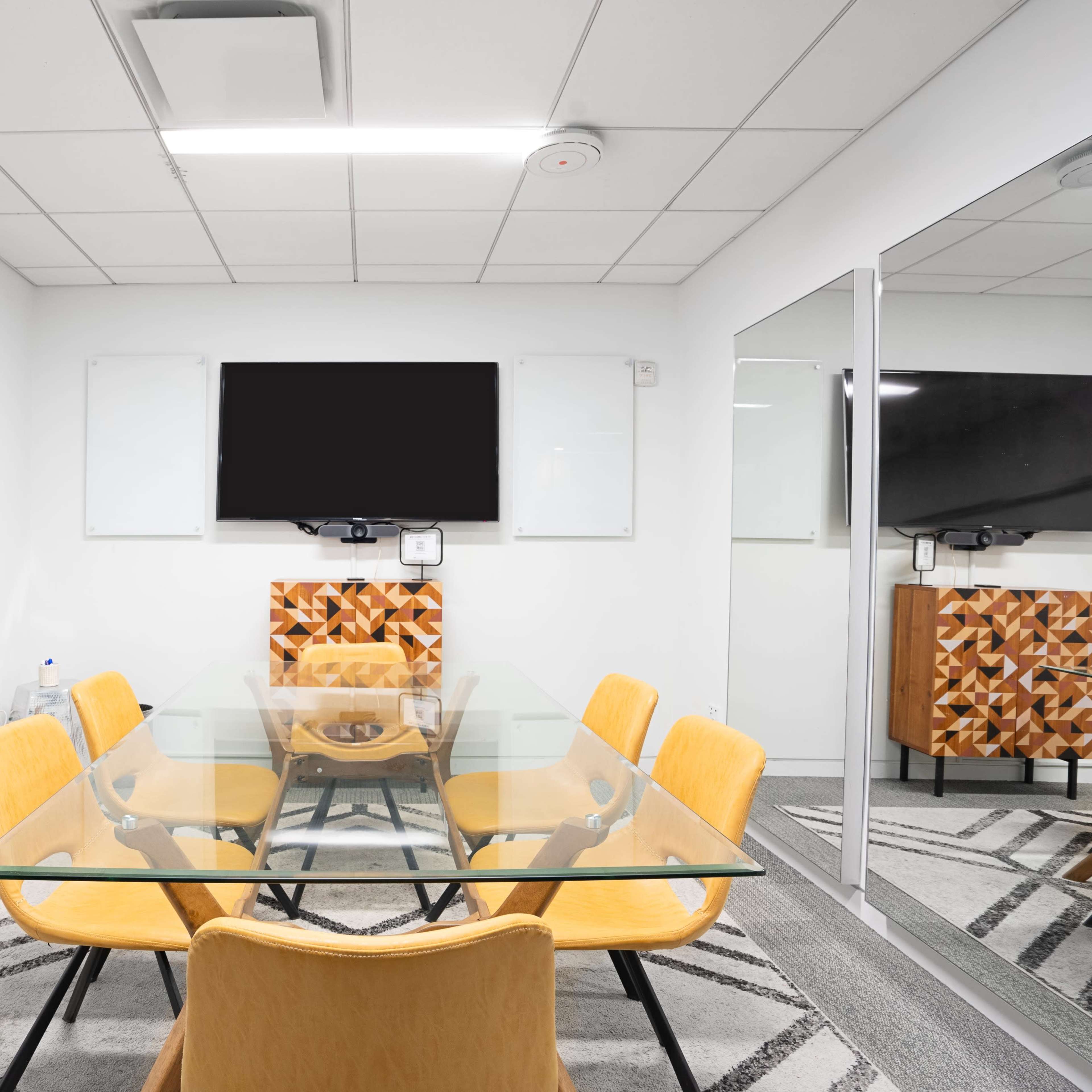 A modern conference room features a glass table surrounded by yellow chairs, a mounted TV, and mirrors on the walls.