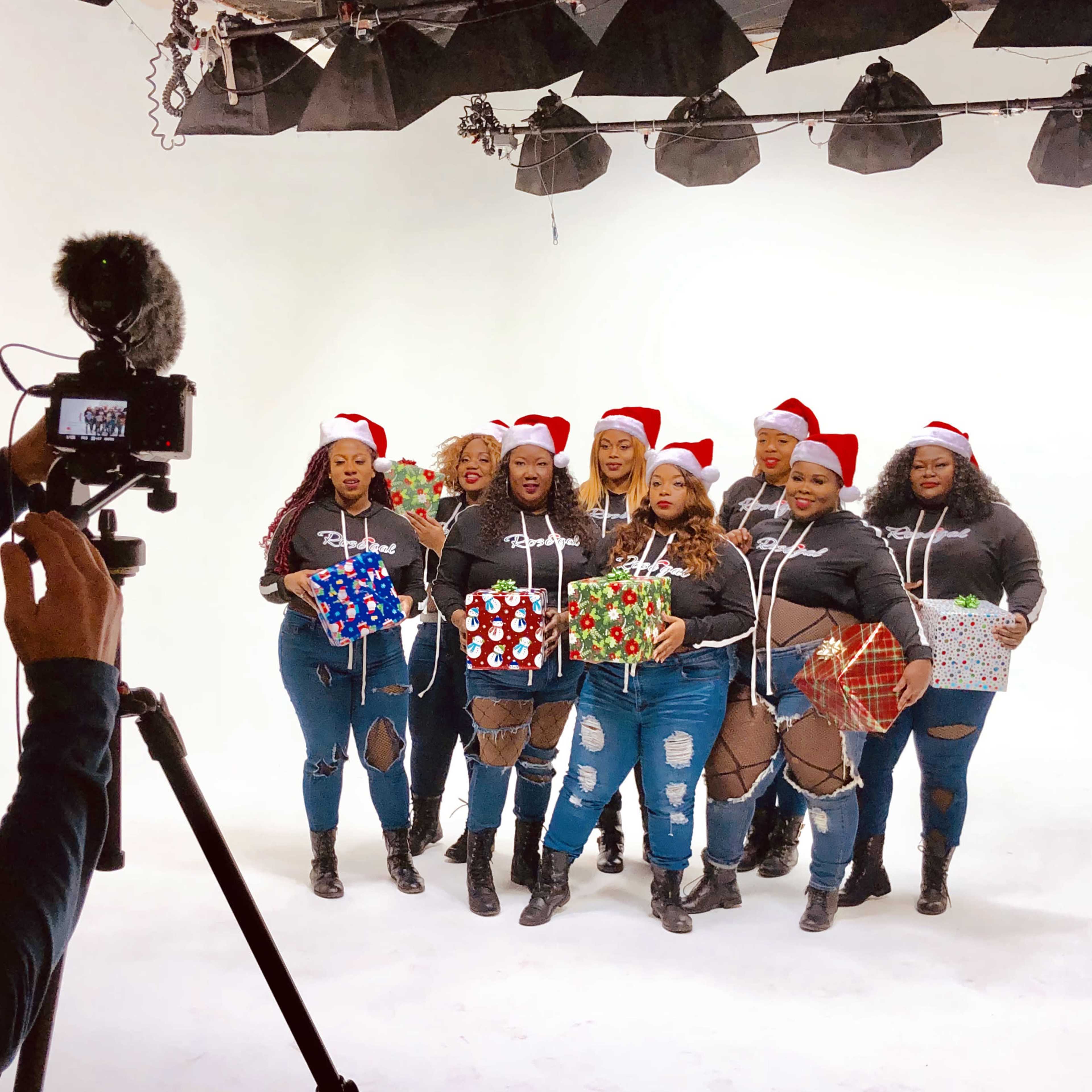 A group of women wearing Santa hats poses with wrapped gifts while being filmed by a person holding a camera.