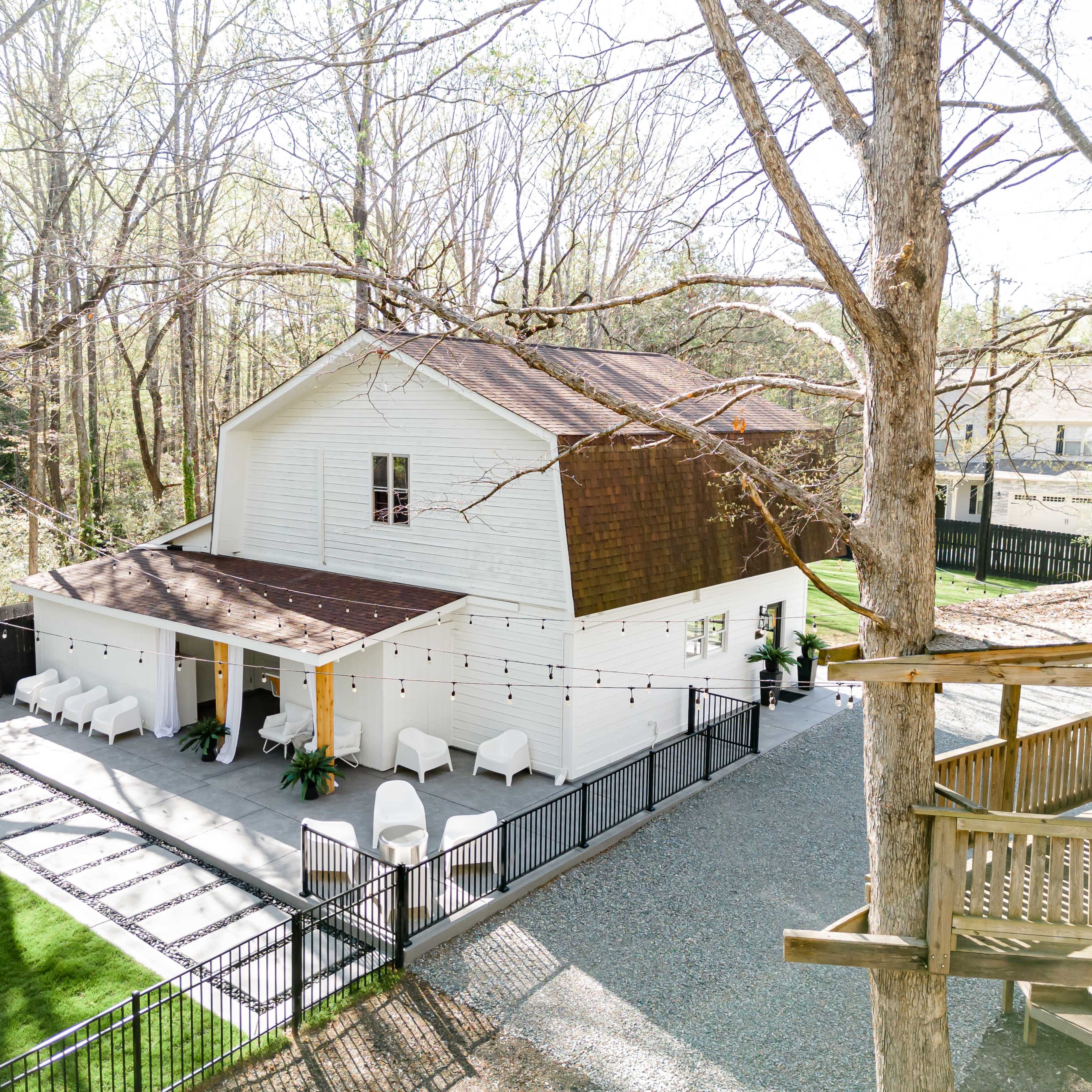 A two-story, white barn-style house features a spacious patio and a fenced yard surrounded by trees.