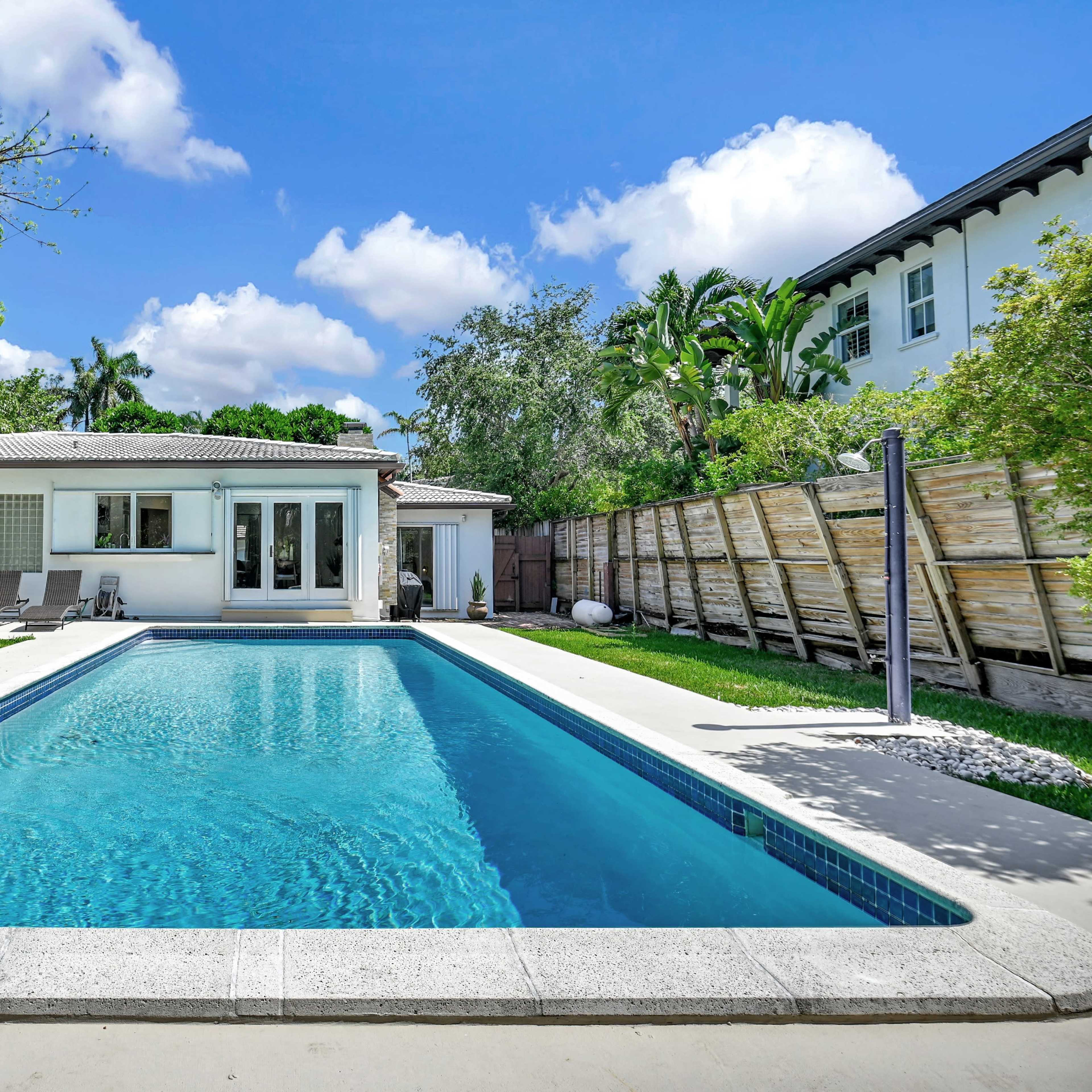 A modern house with a swimming pool and lounge area is surrounded by greenery and palm trees.