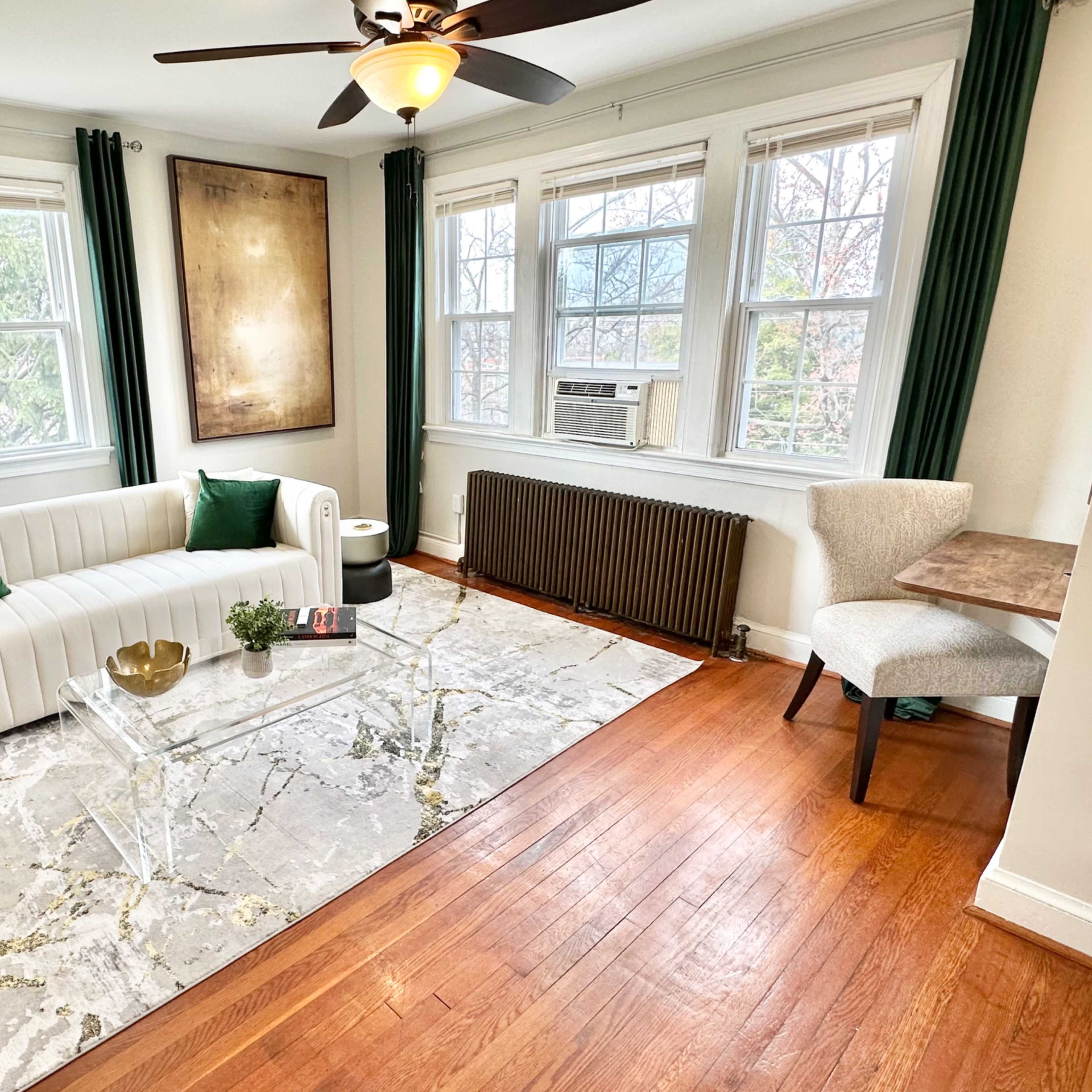 The room features a white sofa with green cushions, a transparent glass coffee table, and a light gray area rug, set against a backdrop of large windows and hardwood flooring.