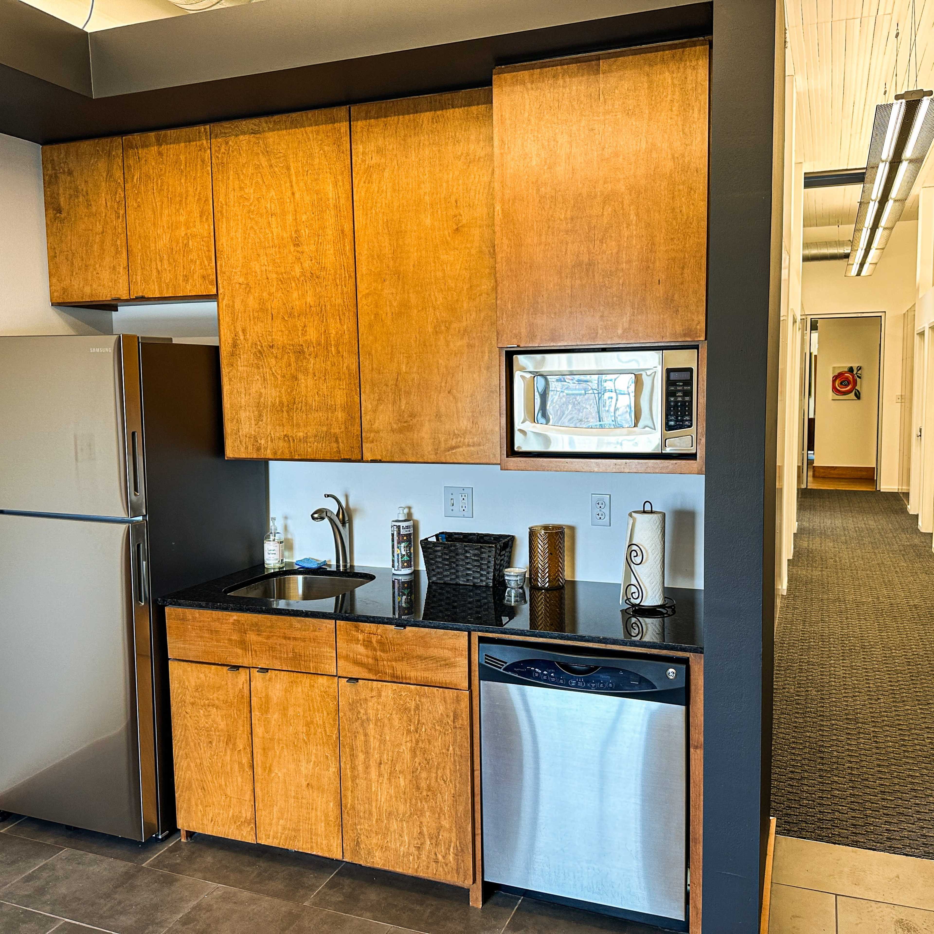 A modern kitchen area with wooden cabinetry, a stainless steel refrigerator, a microwave, and a metal dishwasher, set in a brightly lit space with a carpeted hallway in the background.