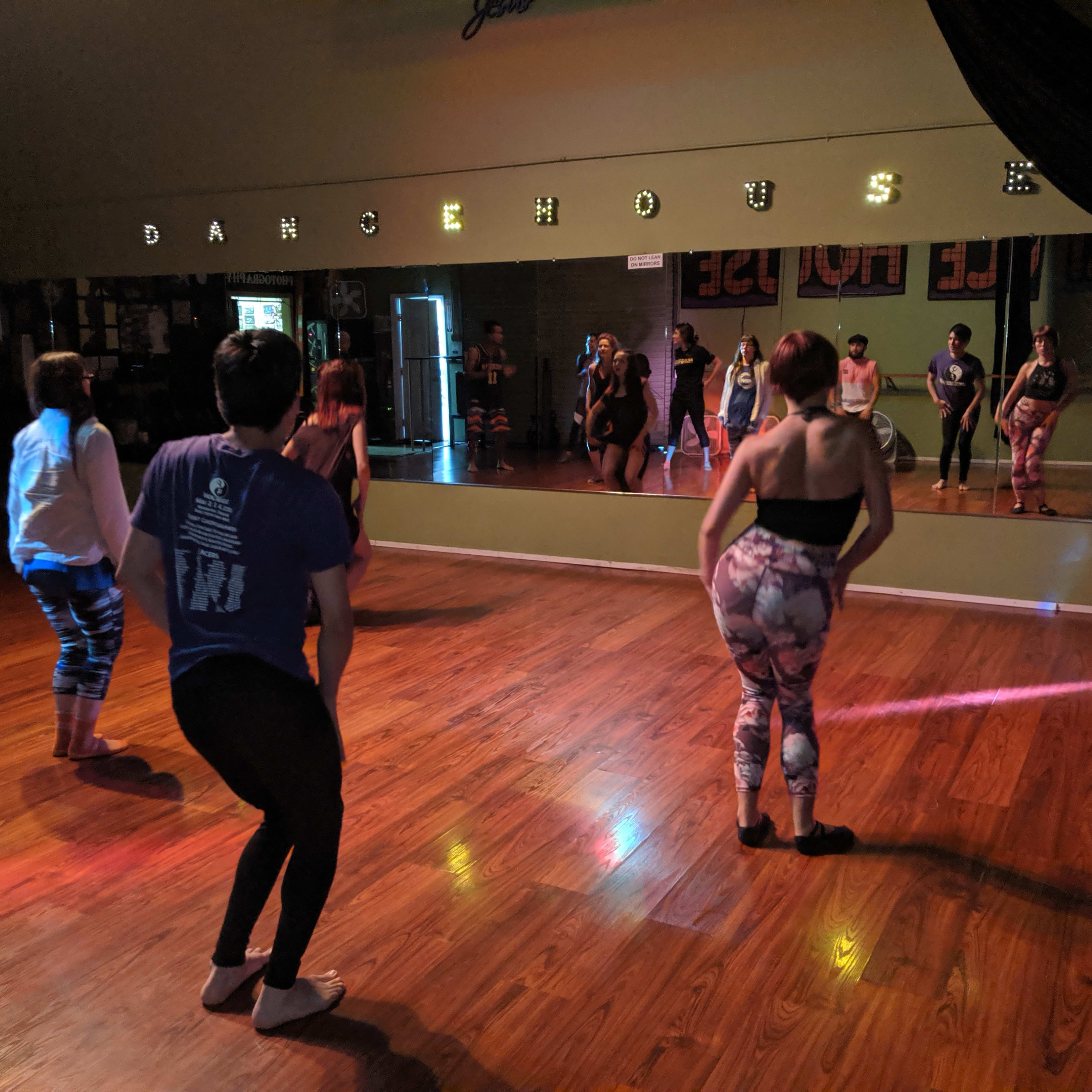 A group of dancers practices in a studio with wooden floors and a large mirror.