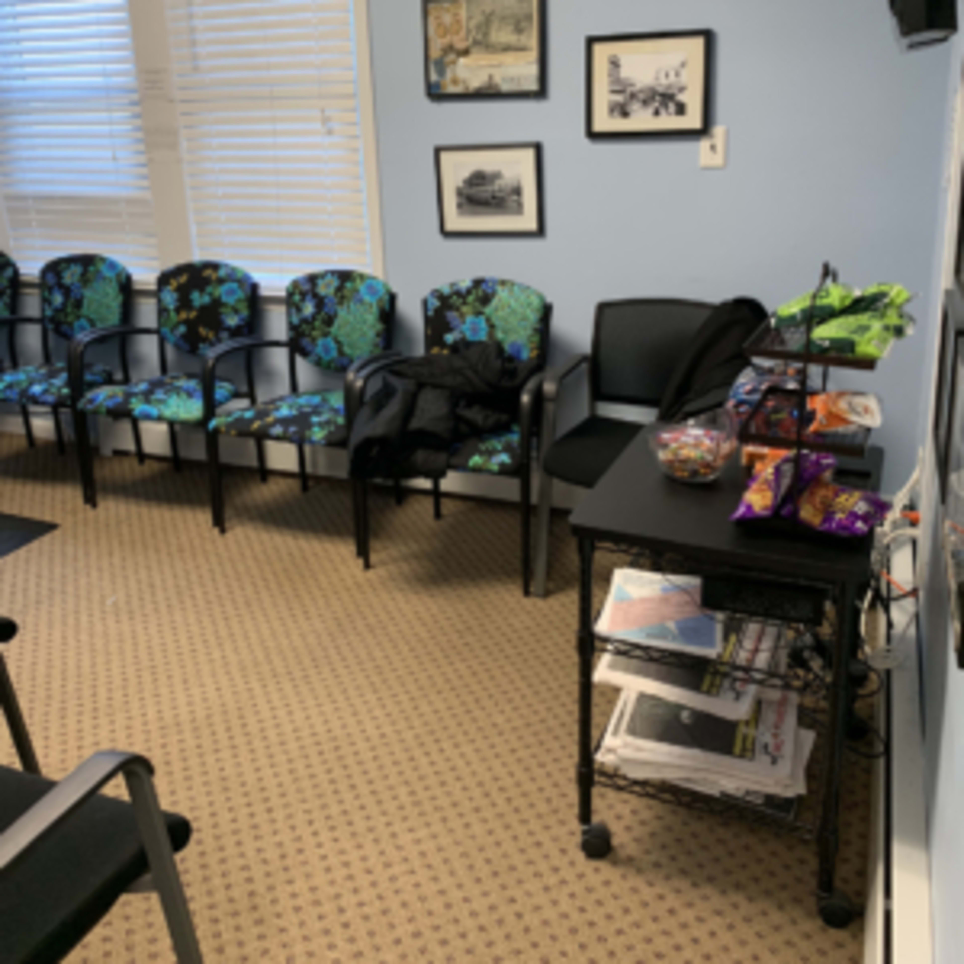 A waiting room with several patterned chairs, a table covered with snacks and magazines, and framed pictures on the wall.