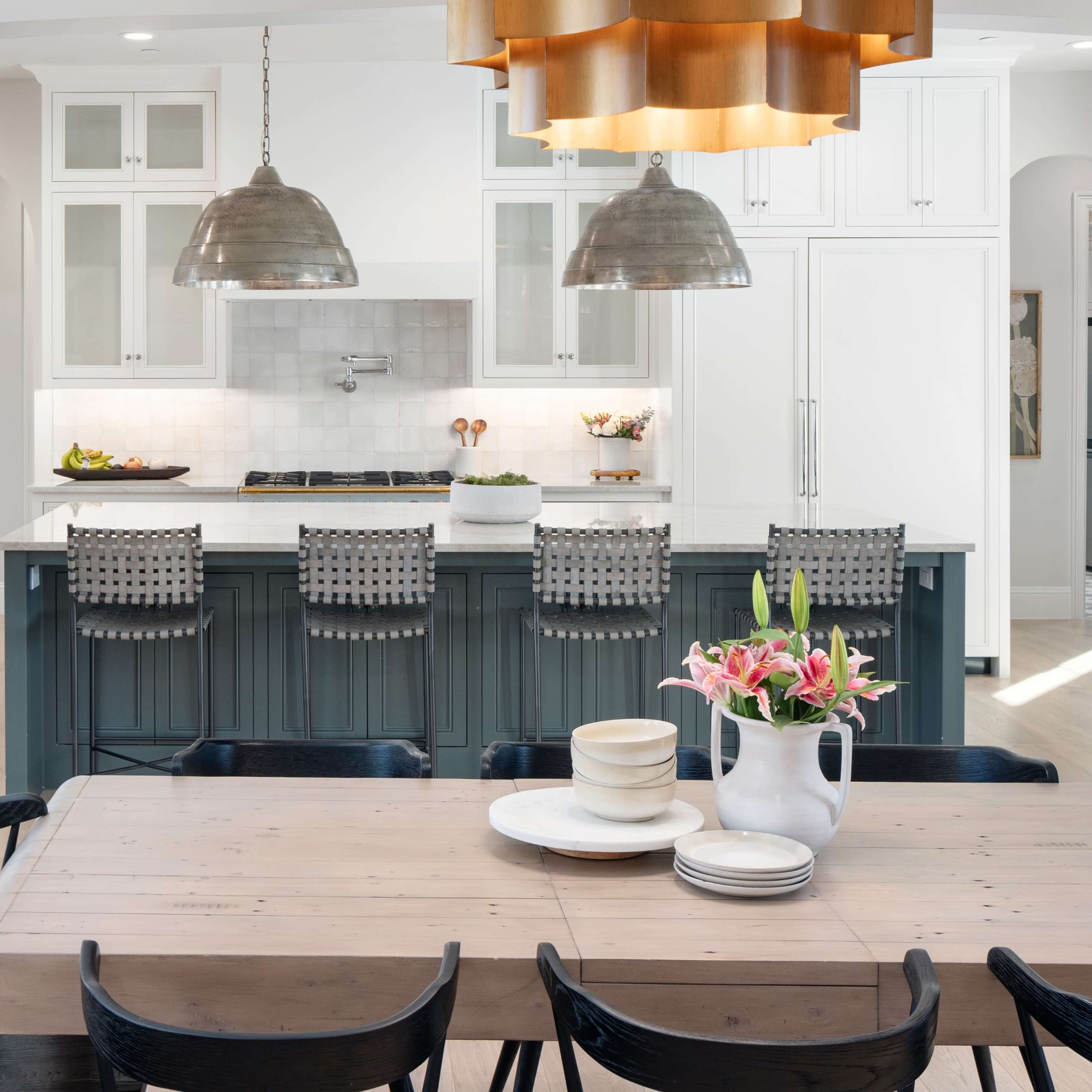 A modern kitchen features an island with bar stools, pendant lights, and a dining table with floral arrangements.