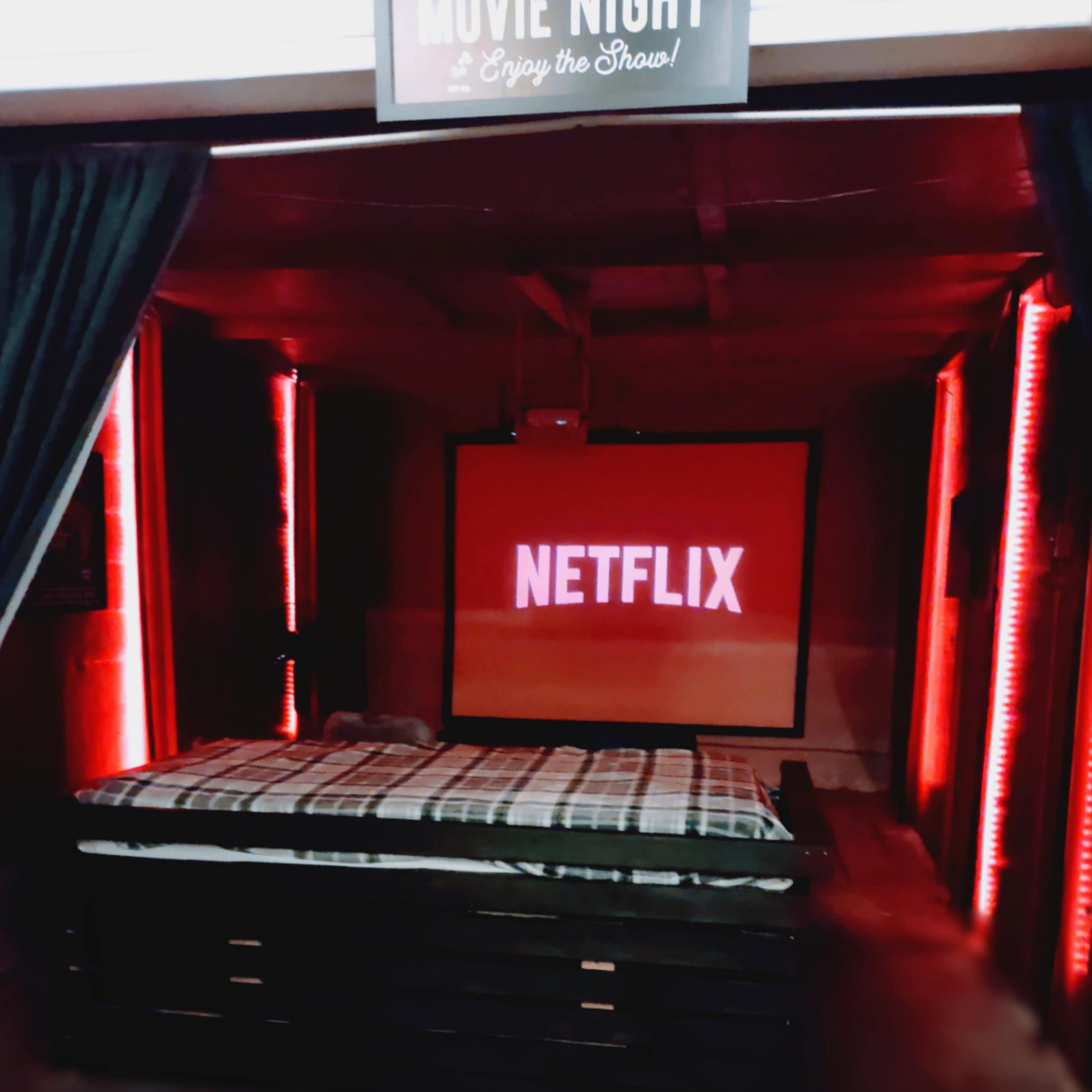 A cozy movie night setup featuring a large screen displaying the Netflix logo, a bed with a checkered blanket, and red LED lights illuminating the space.