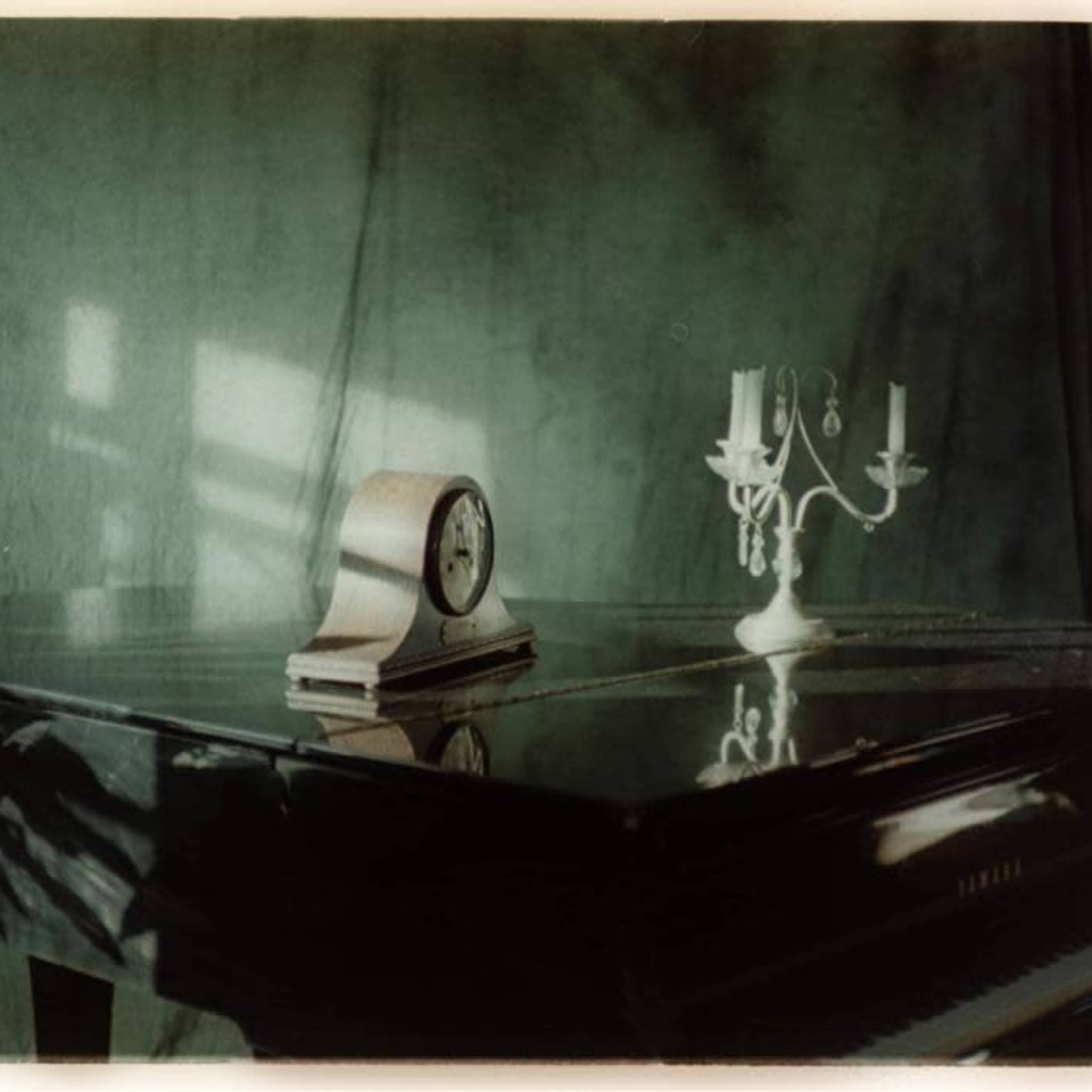 A polished grand piano is positioned with a clock and a candelabrum on its surface, set against a softly lit green backdrop.