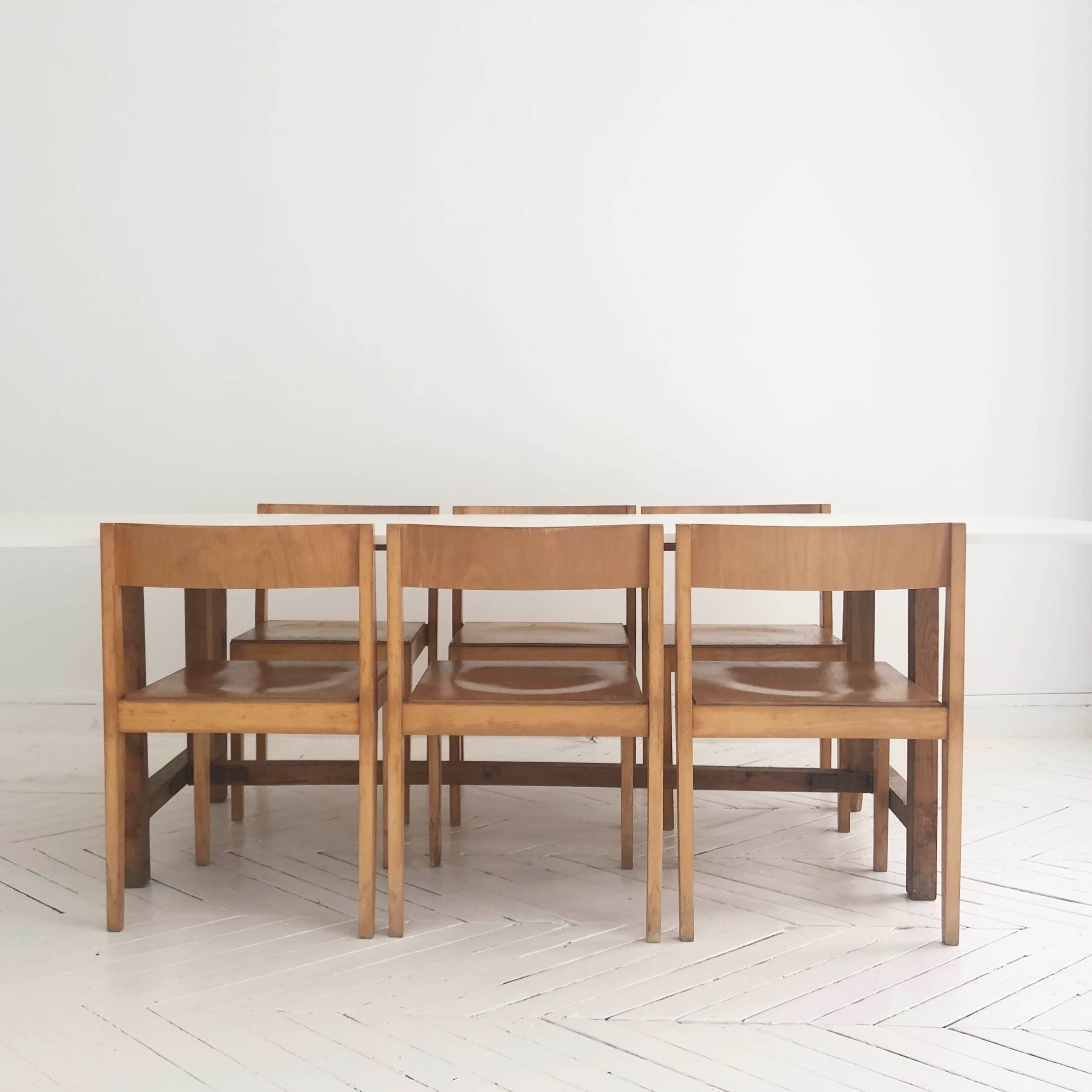 A wooden dining table is flanked by five wooden chairs in a minimalist room with white walls and flooring.