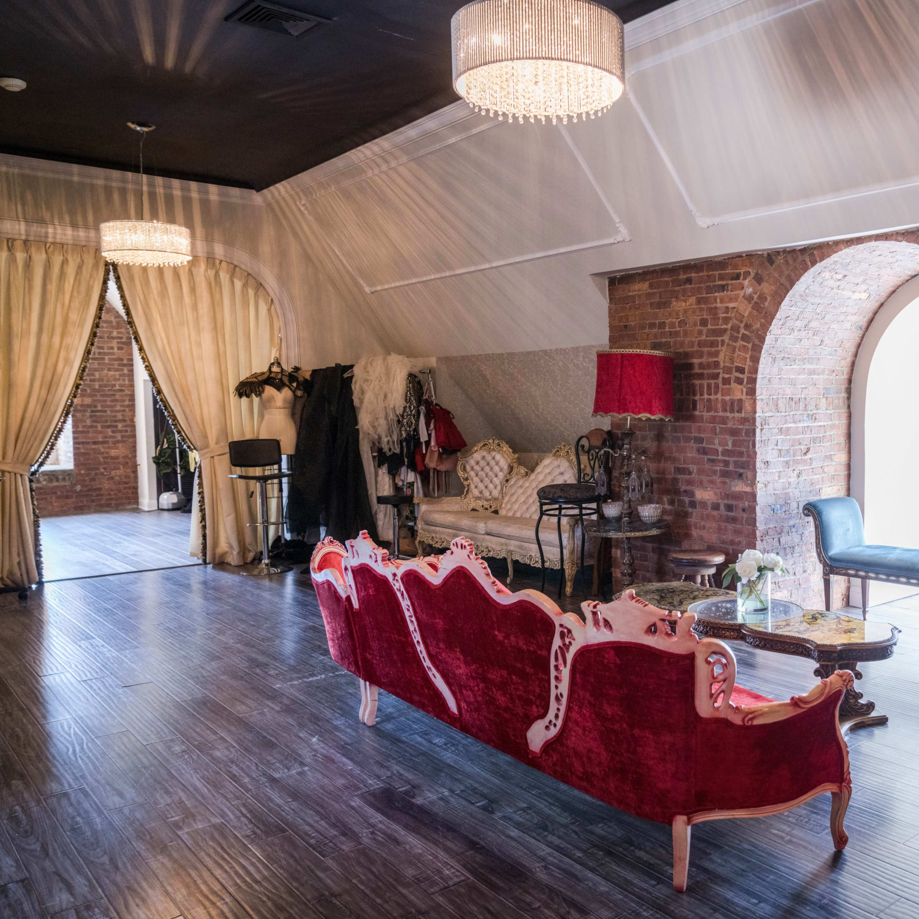 A spacious room with brick walls, elegant vintage furniture, and large arched windows, illuminated by chandeliers.
