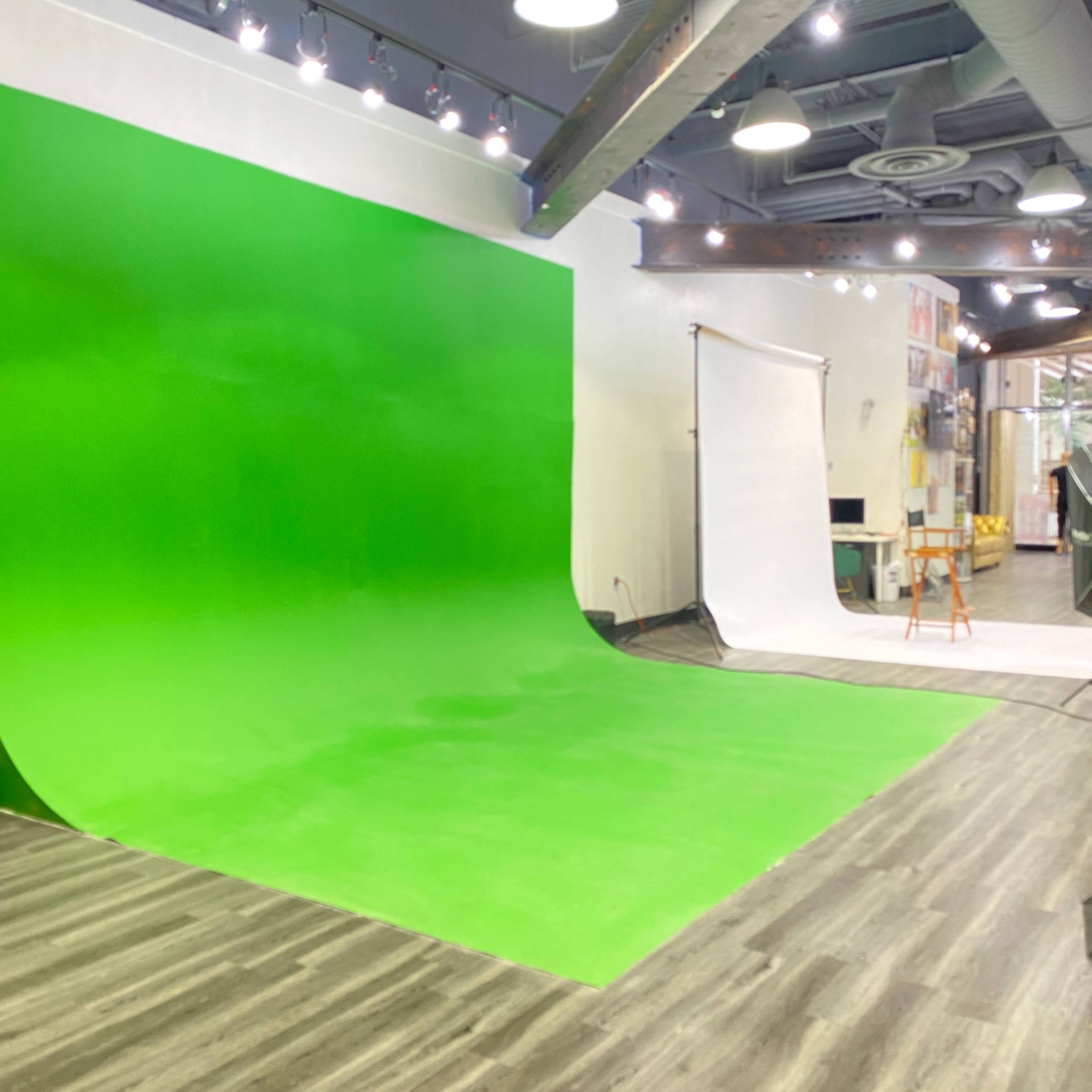 A studio room featuring a large green backdrop and a white backdrop, with wooden flooring and overhead lighting.