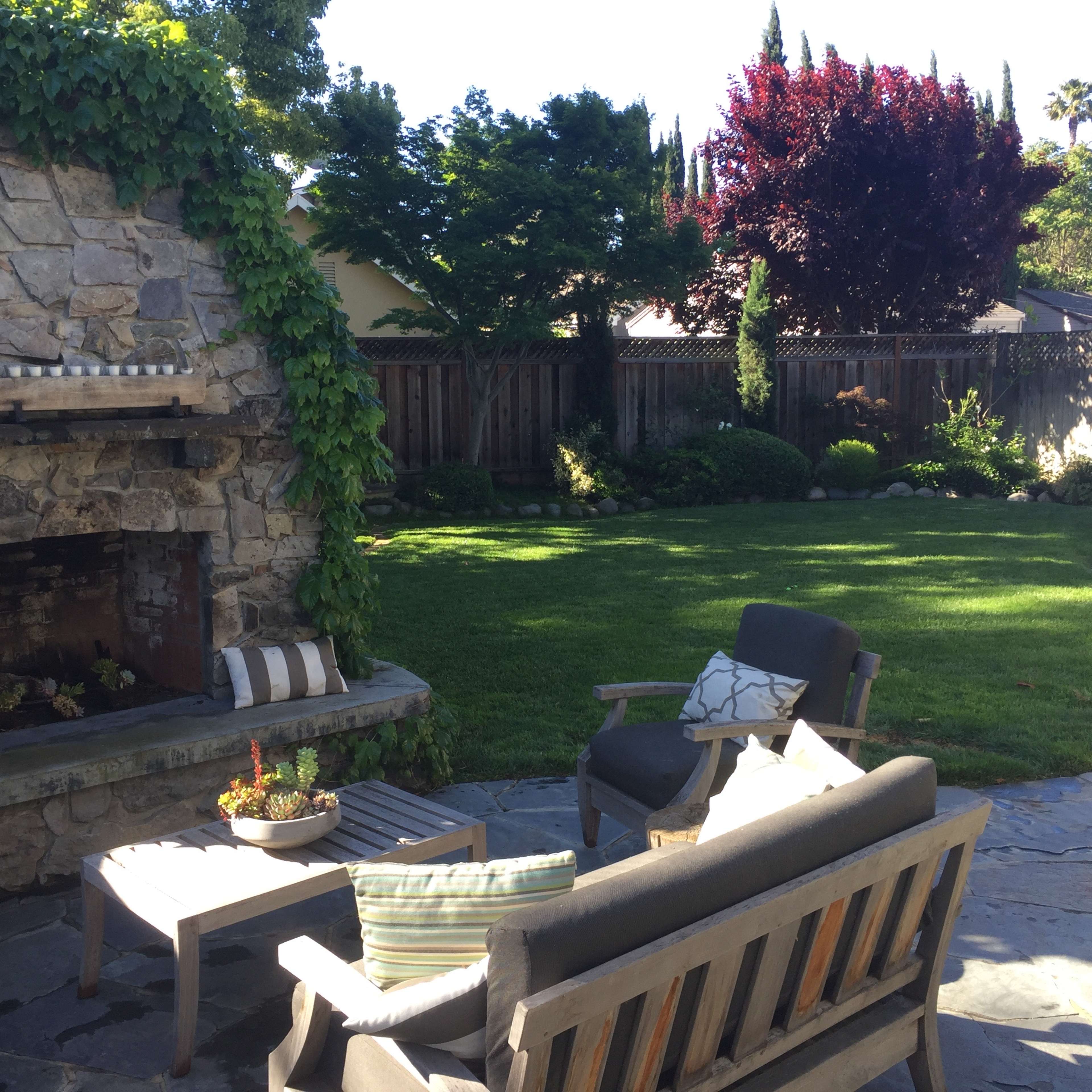 A stone outdoor fireplace is surrounded by a well-maintained lawn and landscaped garden furniture in a residential backyard.