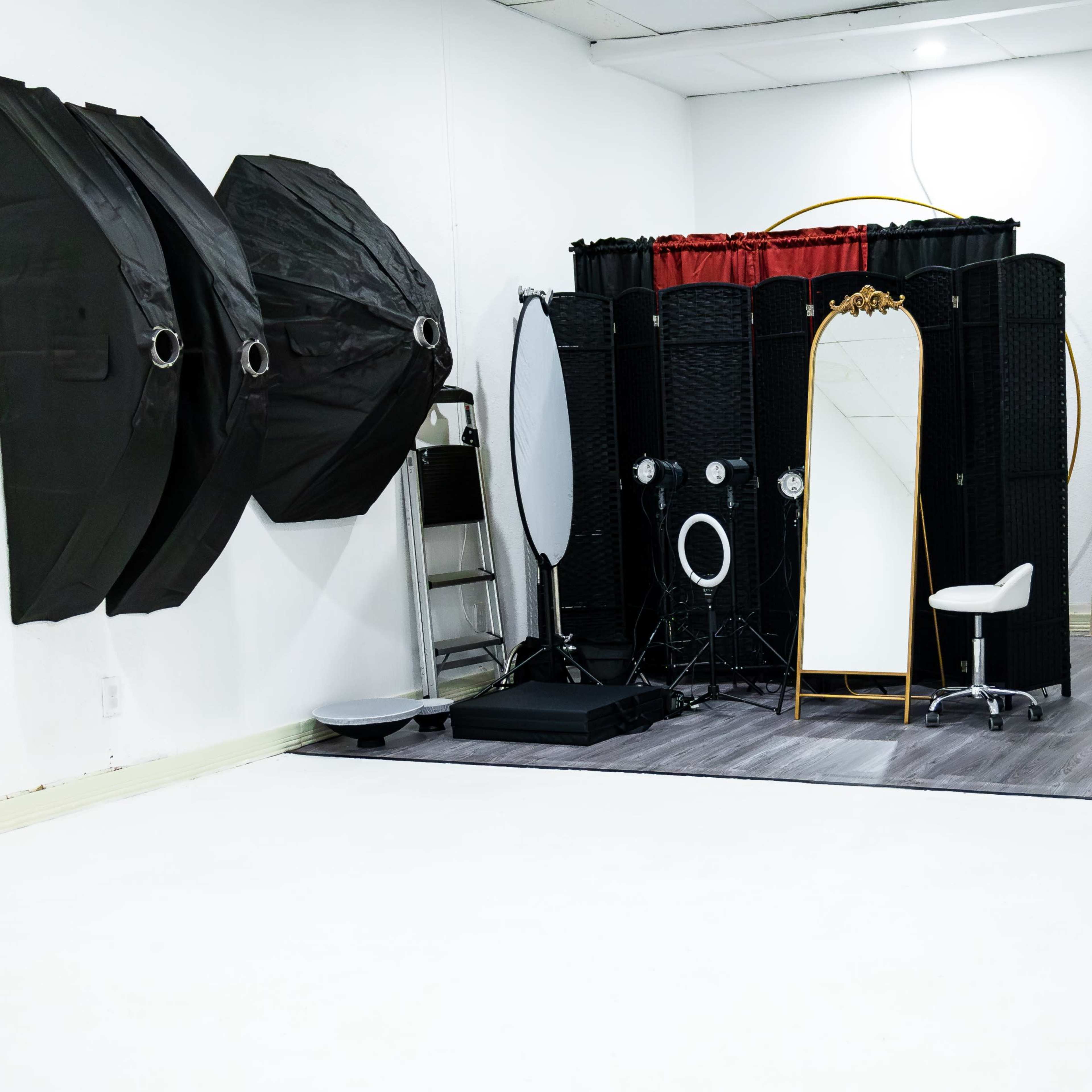 A photography studio featuring black backdrops, a white floor, and a vanity setup with mirrors and lighting equipment.