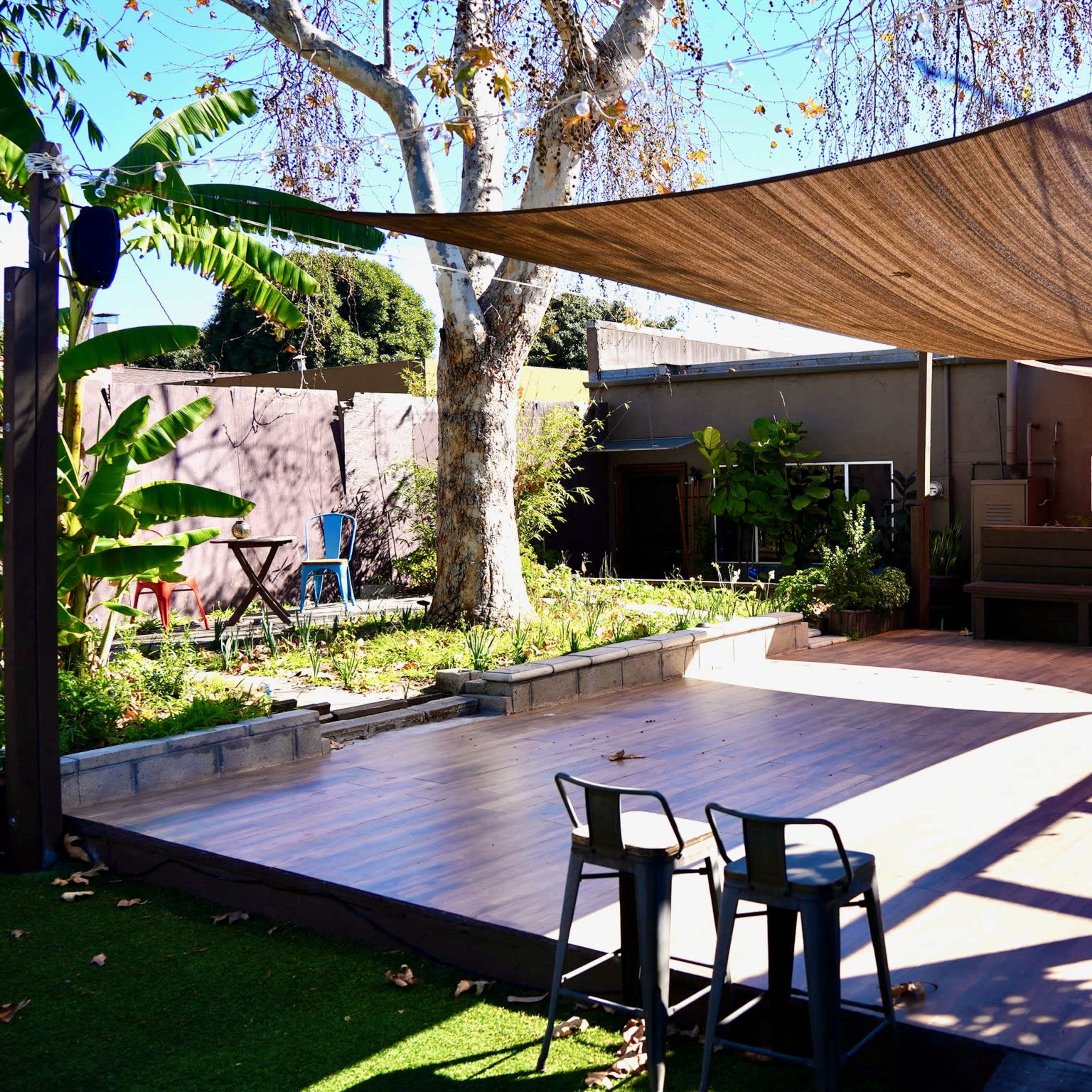 A wooden deck is shaded by a large tarp, surrounded by greenery and a building in the background.