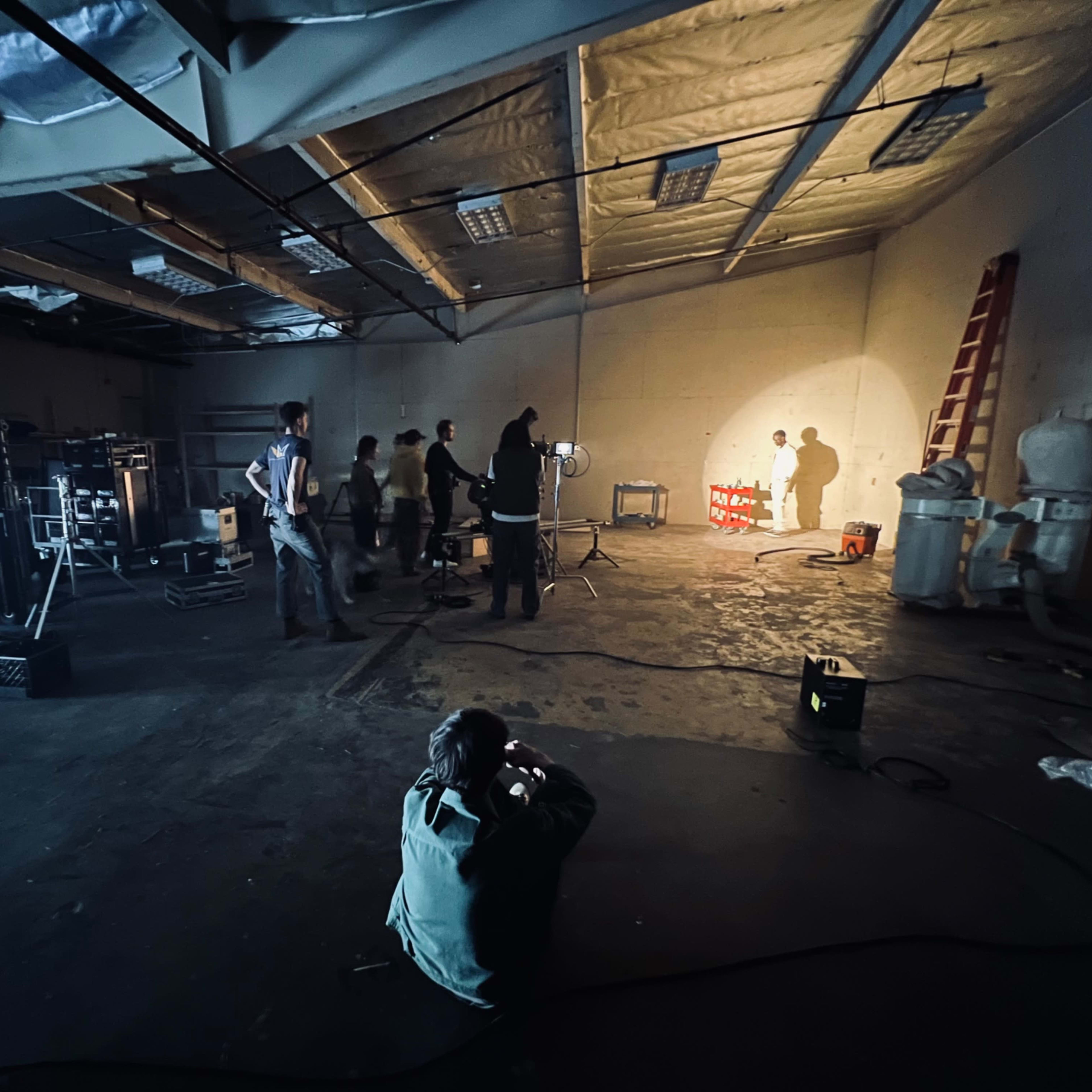 A group of individuals works in an industrial space, using lights and equipment to set up a scene while one person is crouched down photographing.
