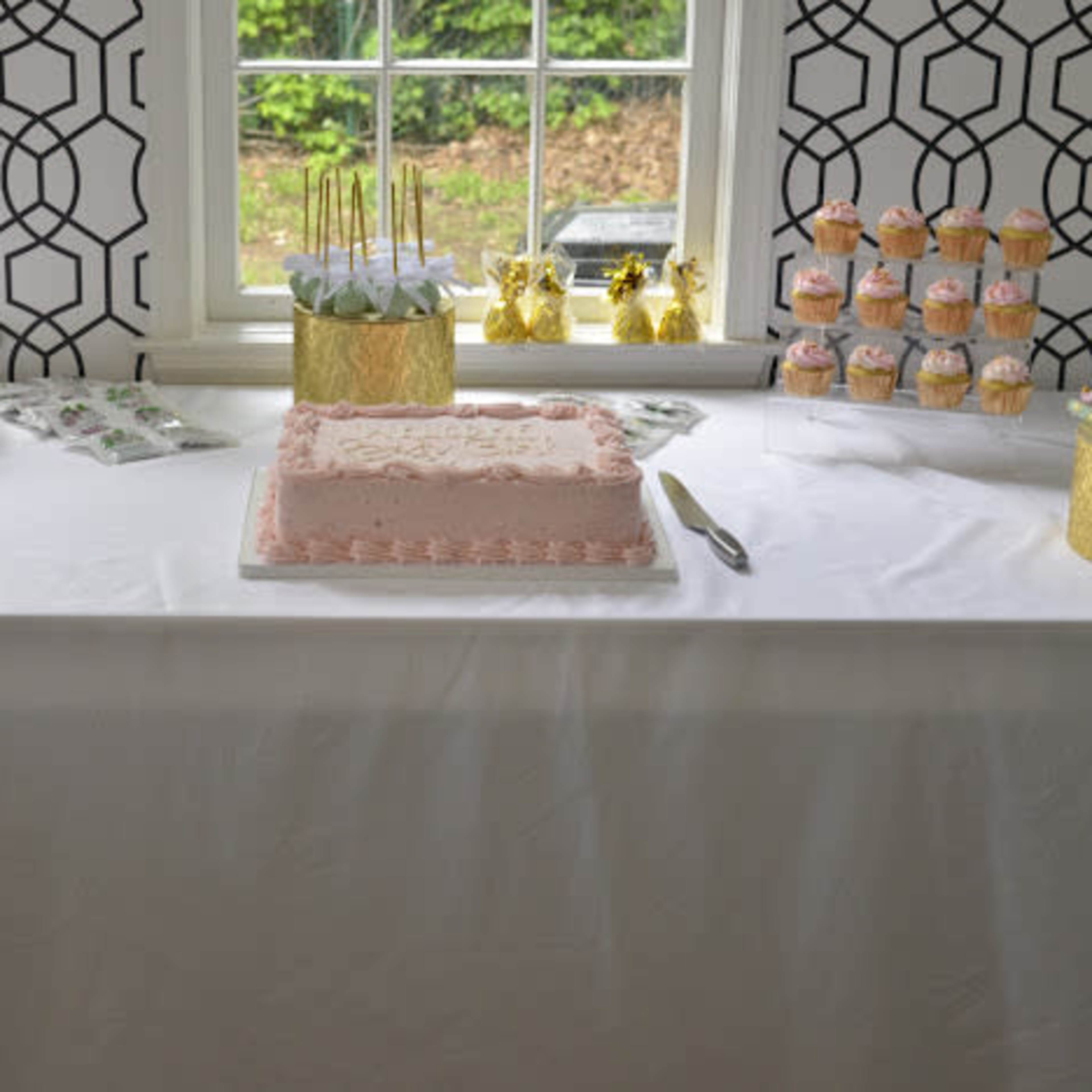 A decorated table features a pink frosted cake at the center, surrounded by trays of cupcakes and dessert treats.