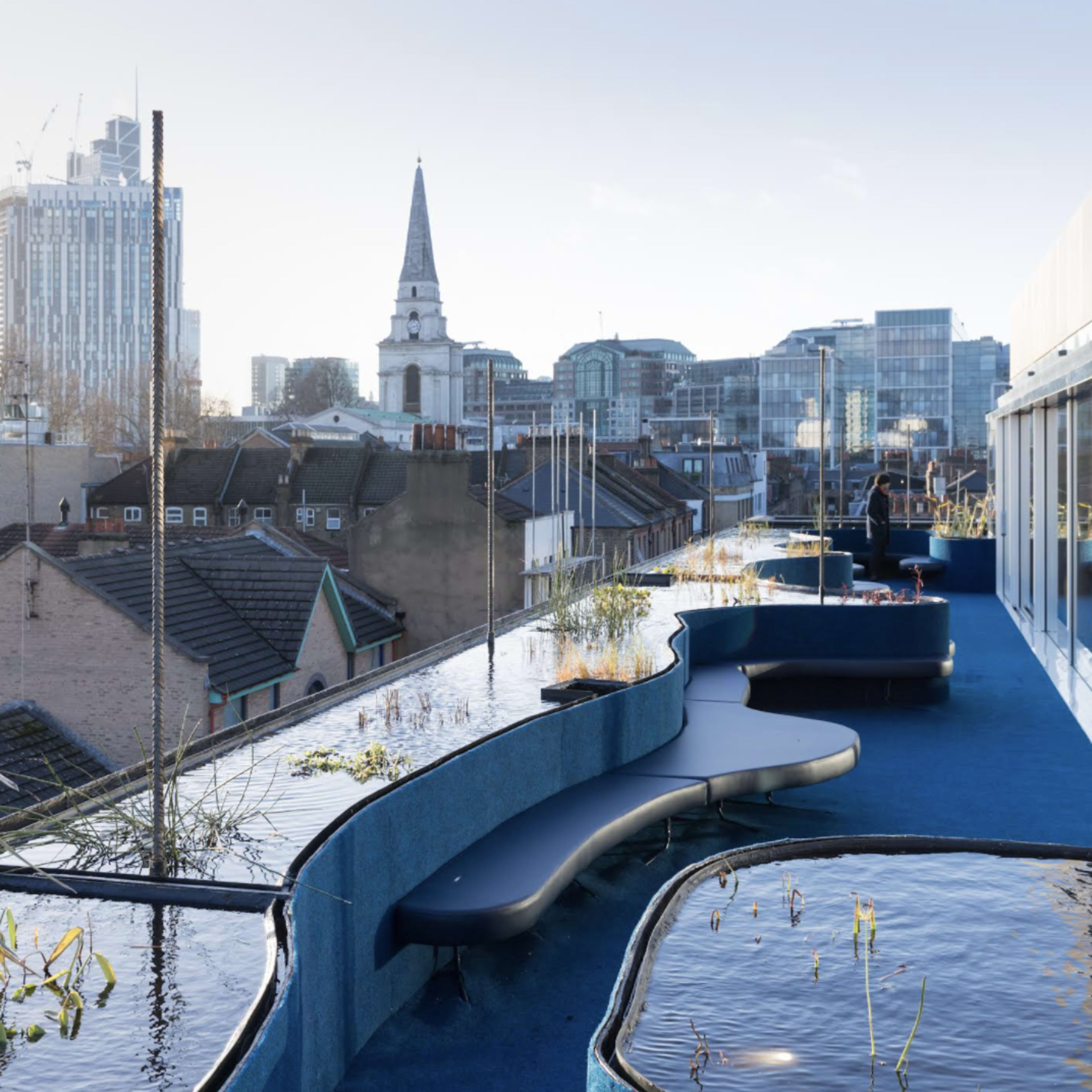 A rooftop terrace with wavy seating along a water feature, overlooking a city skyline with modern buildings and a church spire.