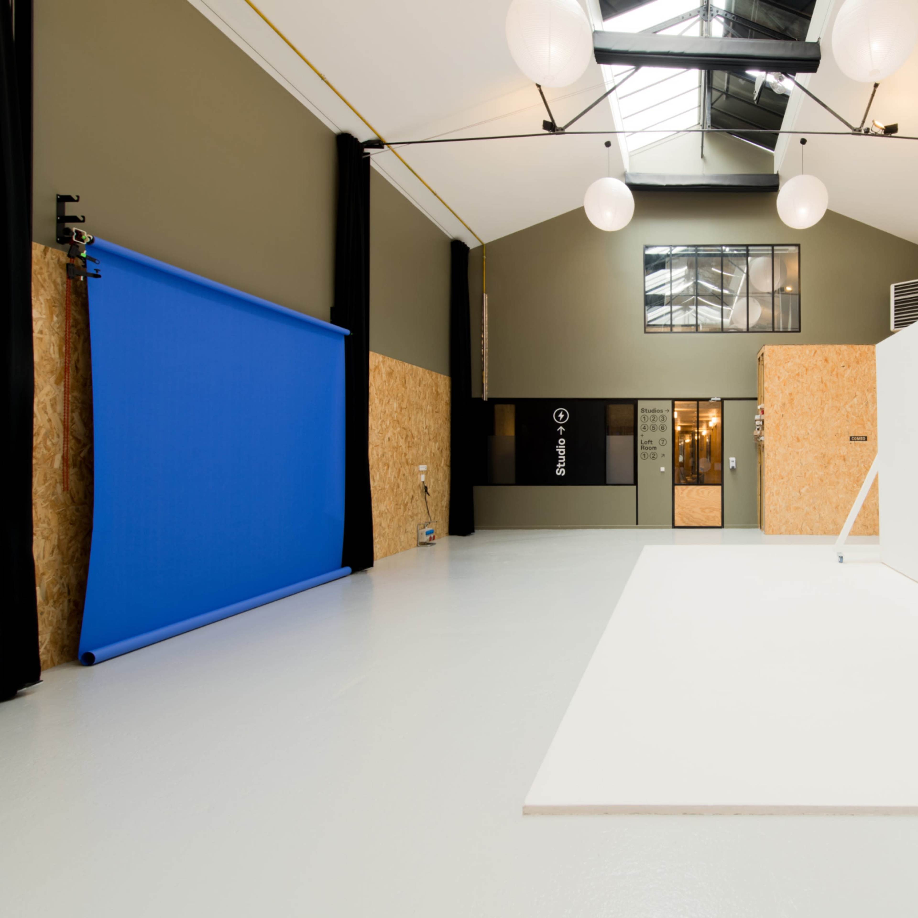 A spacious studio with a blue backdrop on the left, a large white wall, and exposed beams under a skylight.