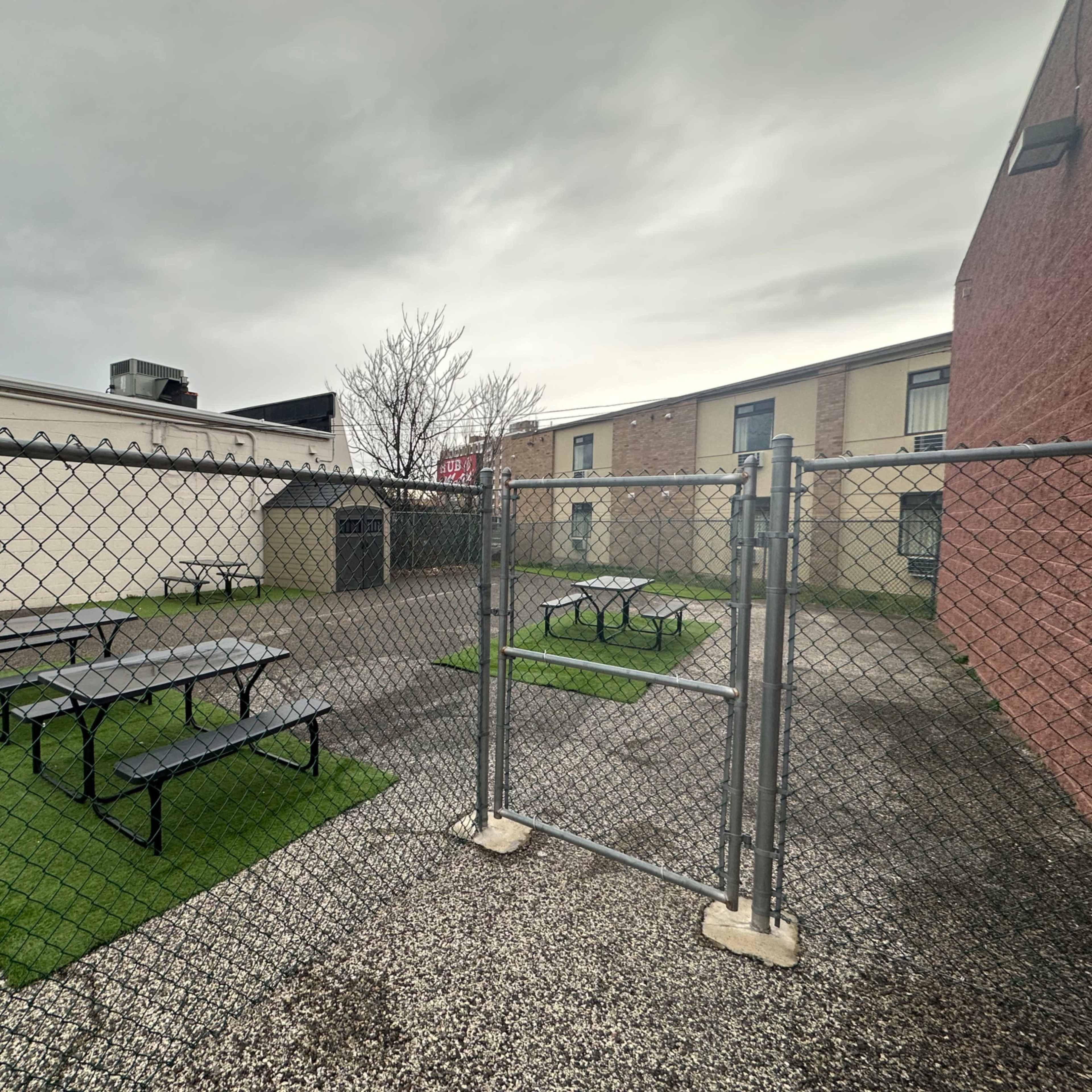 A fenced enclosure with two sets of picnic tables on artificial grass, flanked by buildings under a cloudy sky.