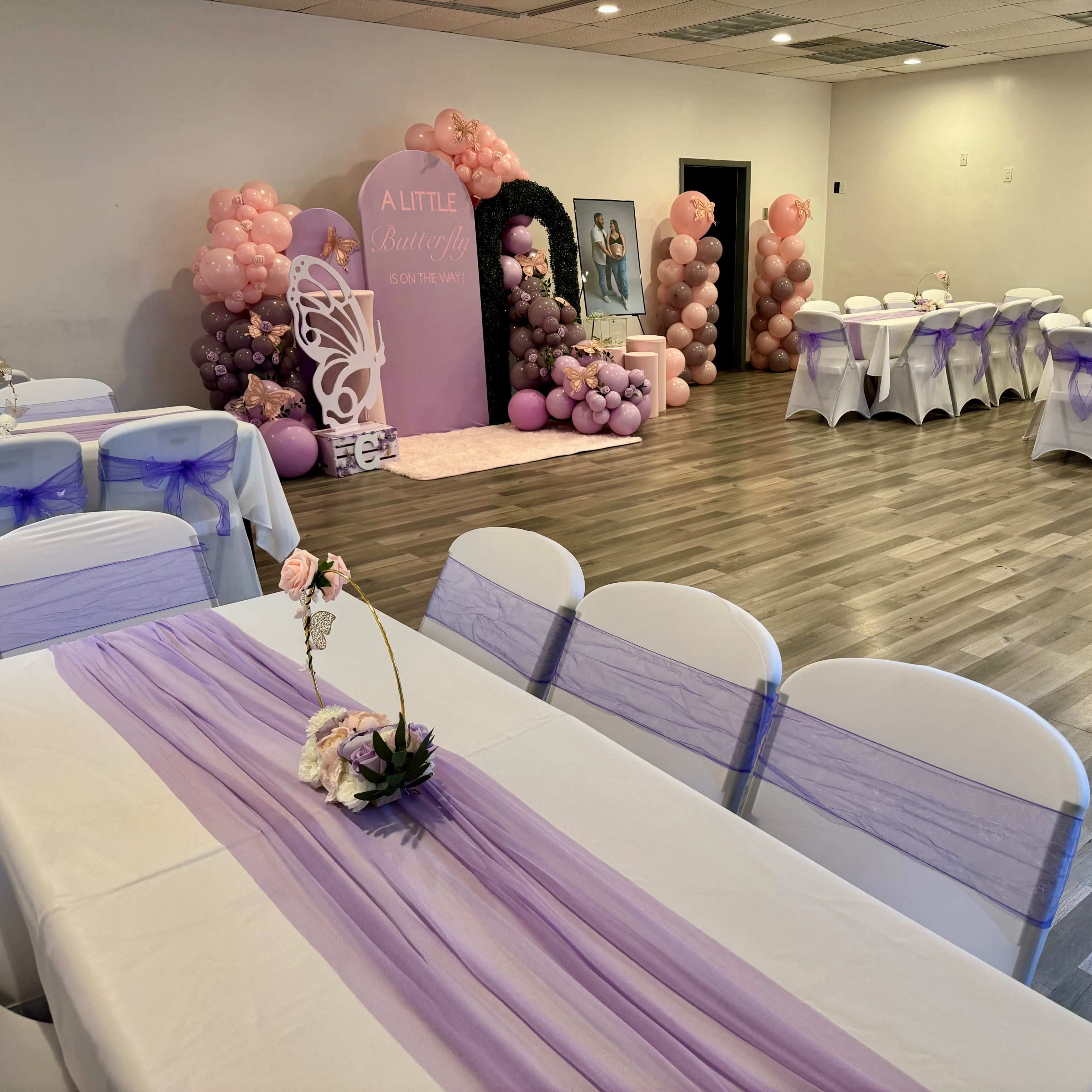 A decorated event space featuring white tables with purple table runners, surrounded by balloon arrangements and a backdrop for a celebration.