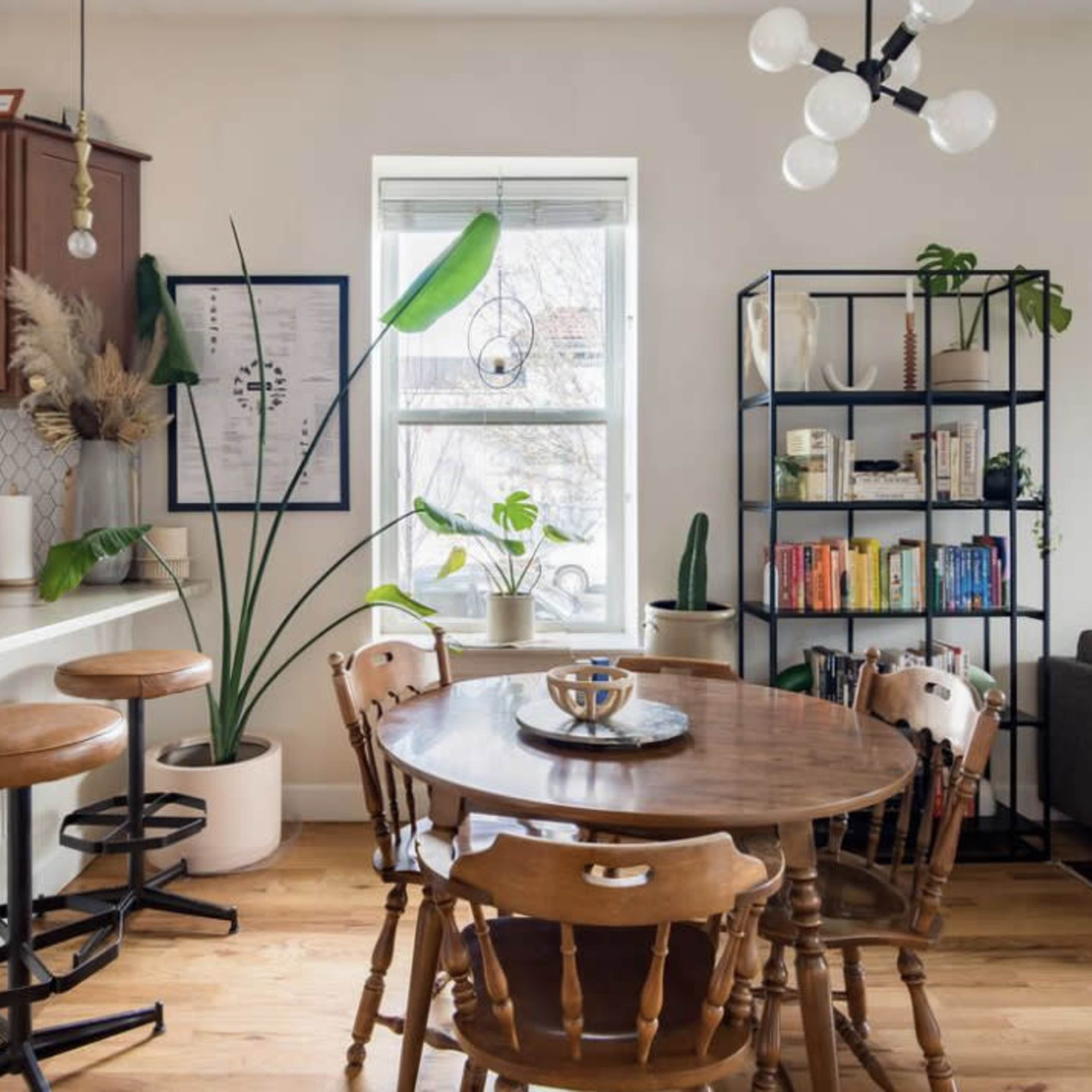A modern dining area with a round wooden table surrounded by chairs, a kitchen bar with stools, and a bookshelf filled with plants and books.