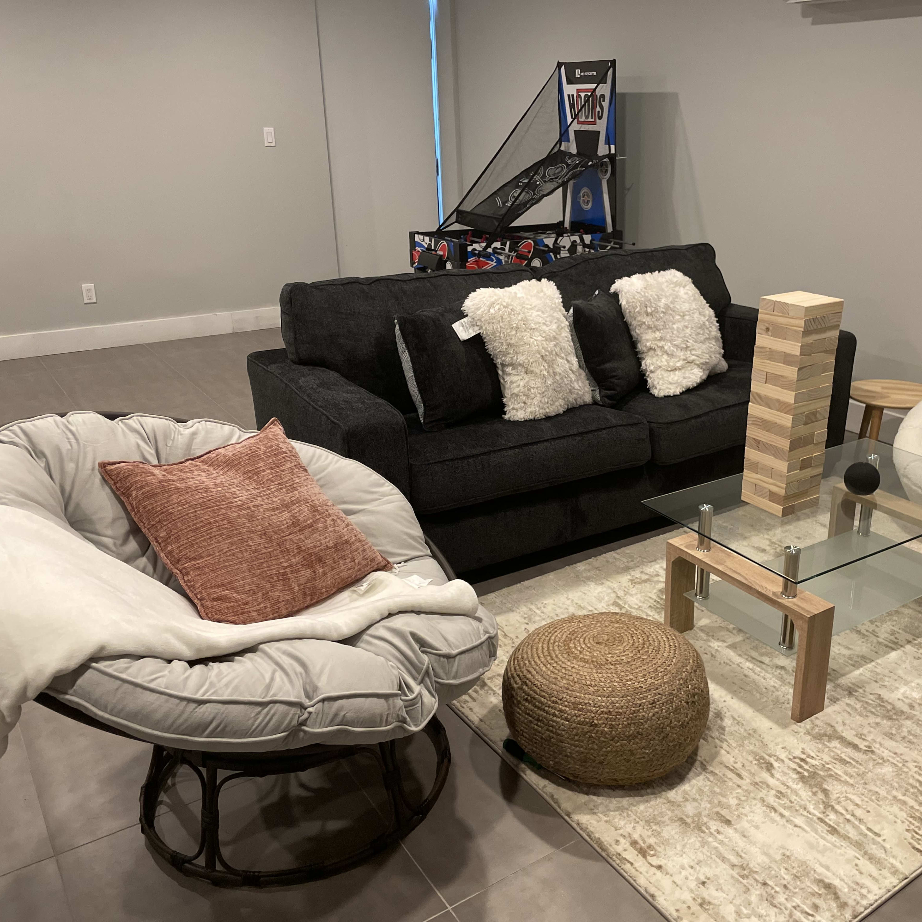 A cozy living area with a black couch, two unique chairs, a glass coffee table, and a decorative rug on a tiled floor.