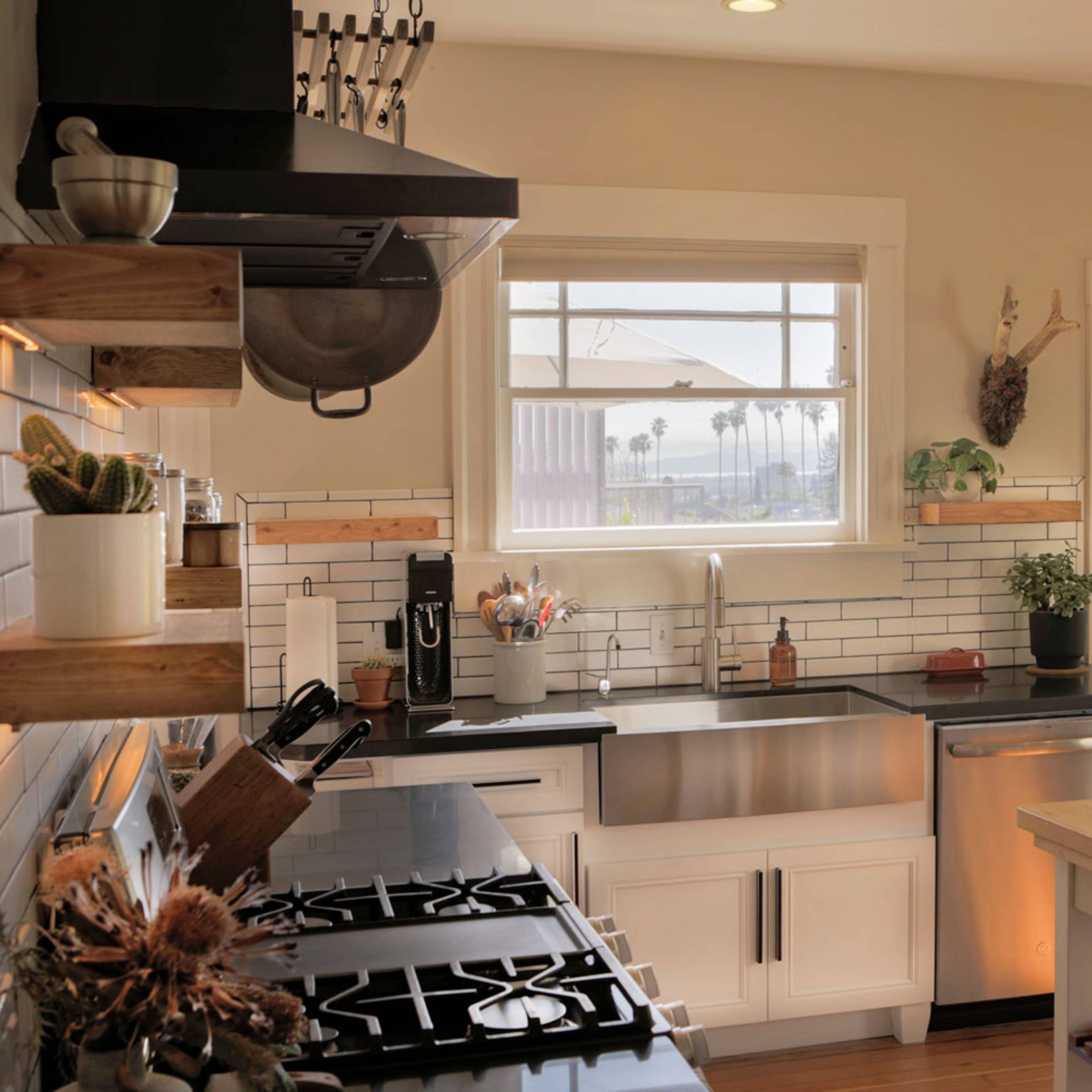 A modern kitchen features stainless steel appliances, a large window overlooking greenery, and wooden shelves displaying kitchenware.
