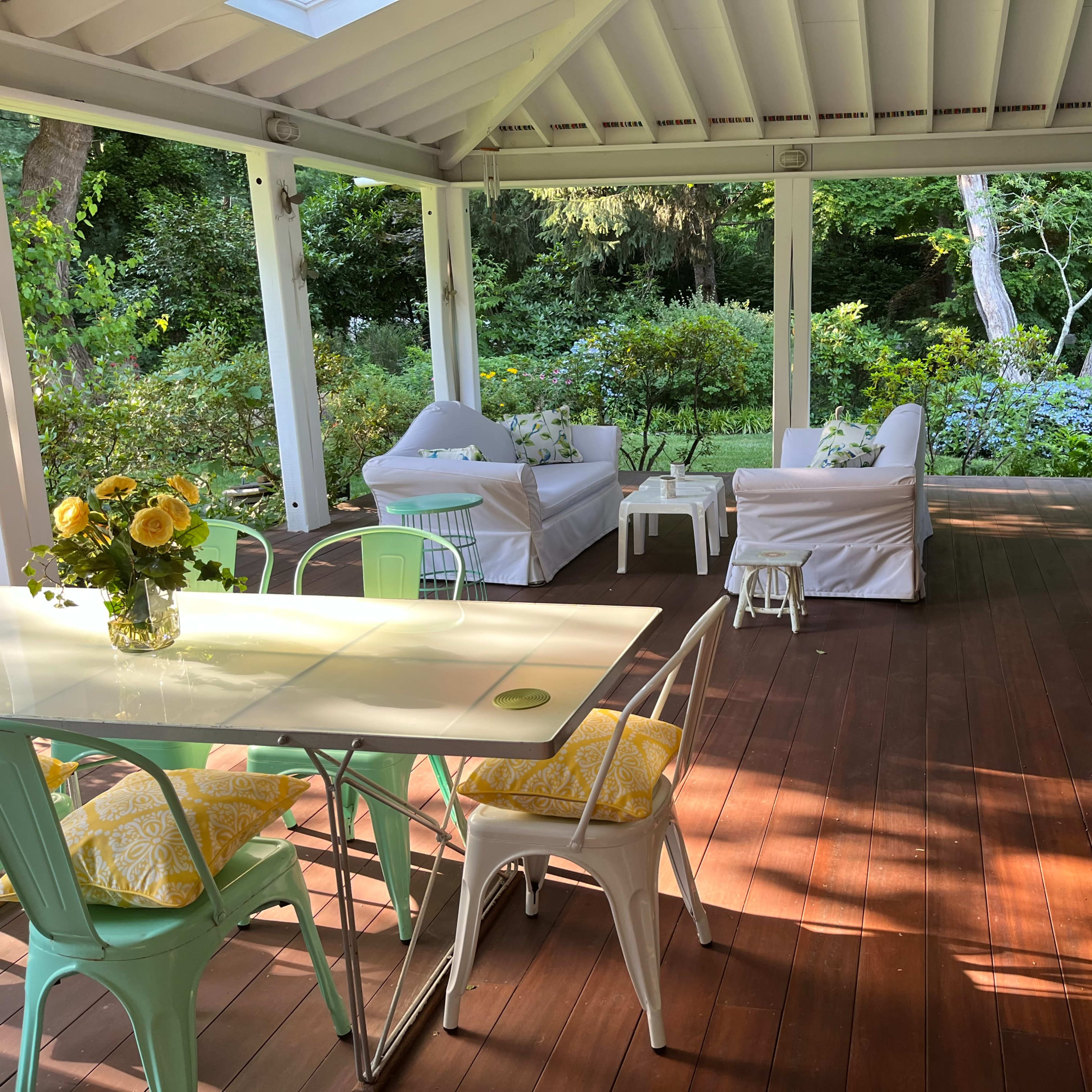 A covered wooden patio features a dining table with colorful chairs and a seating area with white couches surrounded by greenery.