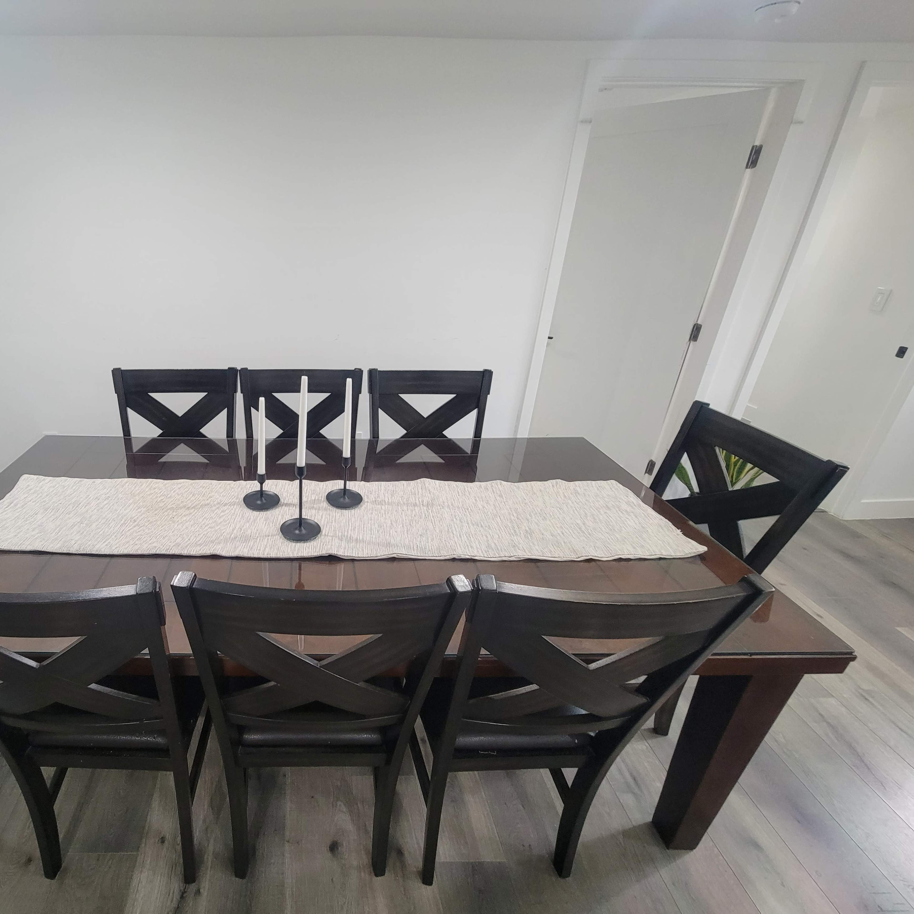A rectangular dining table with a beige runner and four black chairs is positioned in a well-lit room with white walls and wooden flooring.