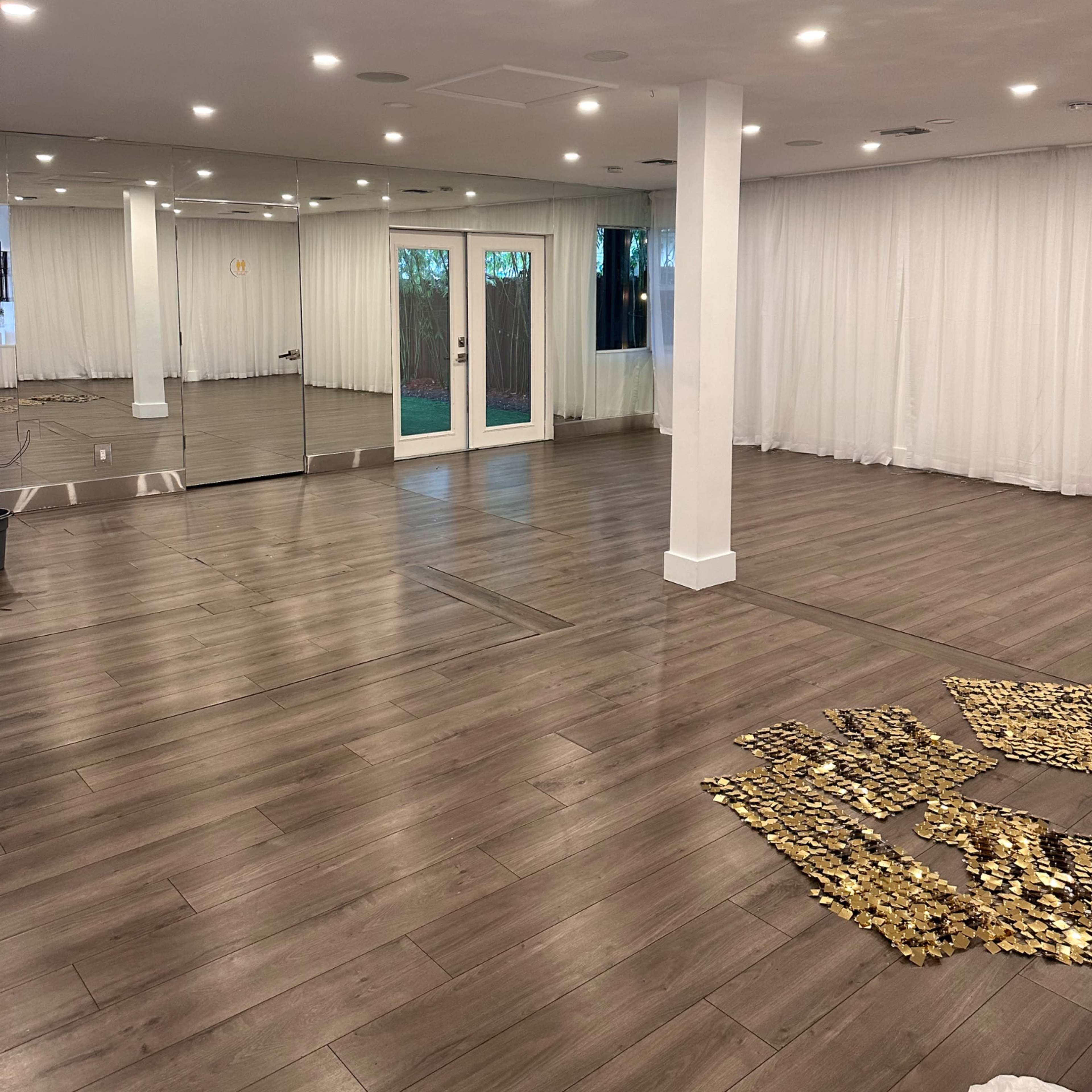 The room features a mirrored wall, large windows with sheer curtains, and a wooden floor, with a pattern arranged on the floor.