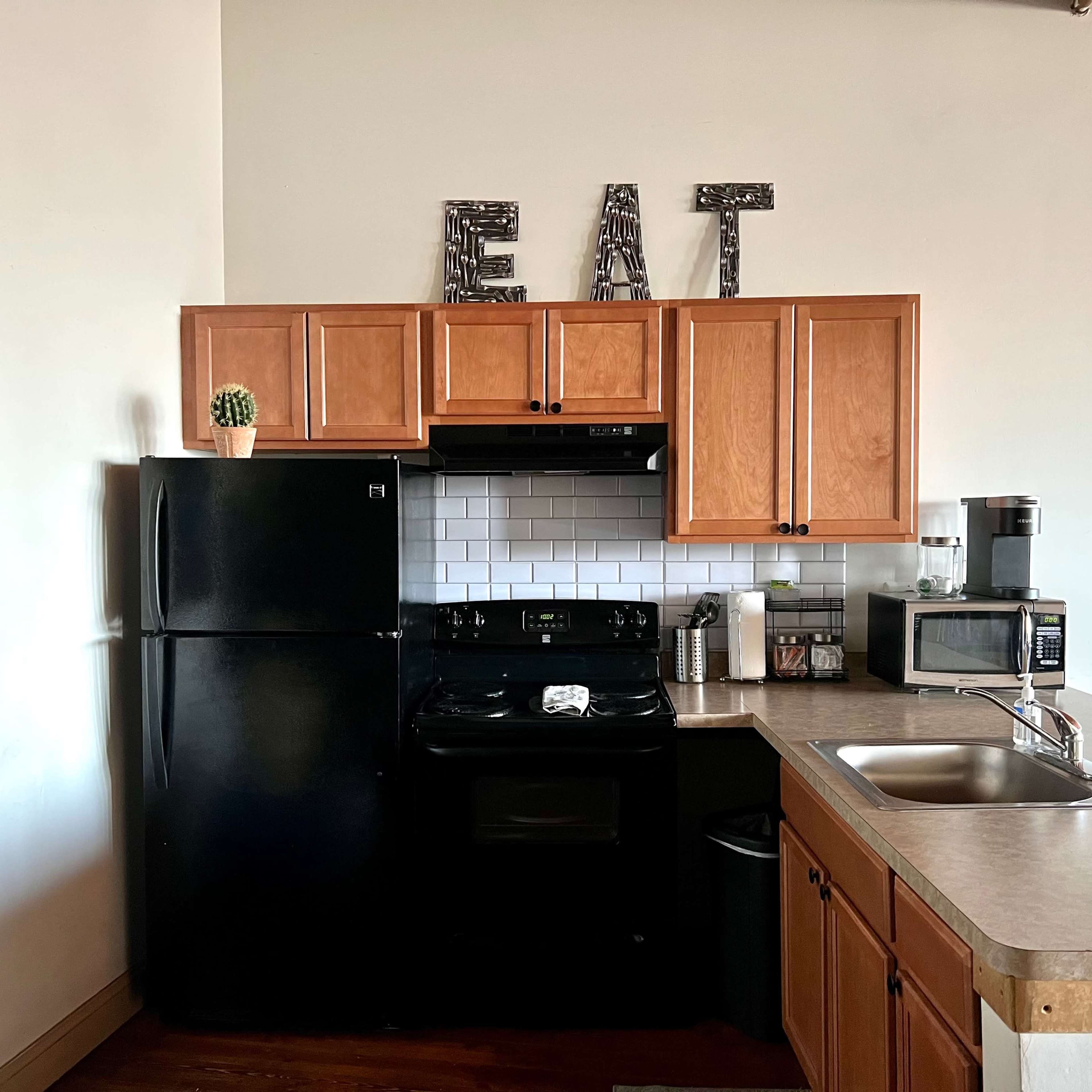 A kitchen features wooden cabinets, a black refrigerator, a stove, a microwave, and the word "EAT" displayed above the countertop.