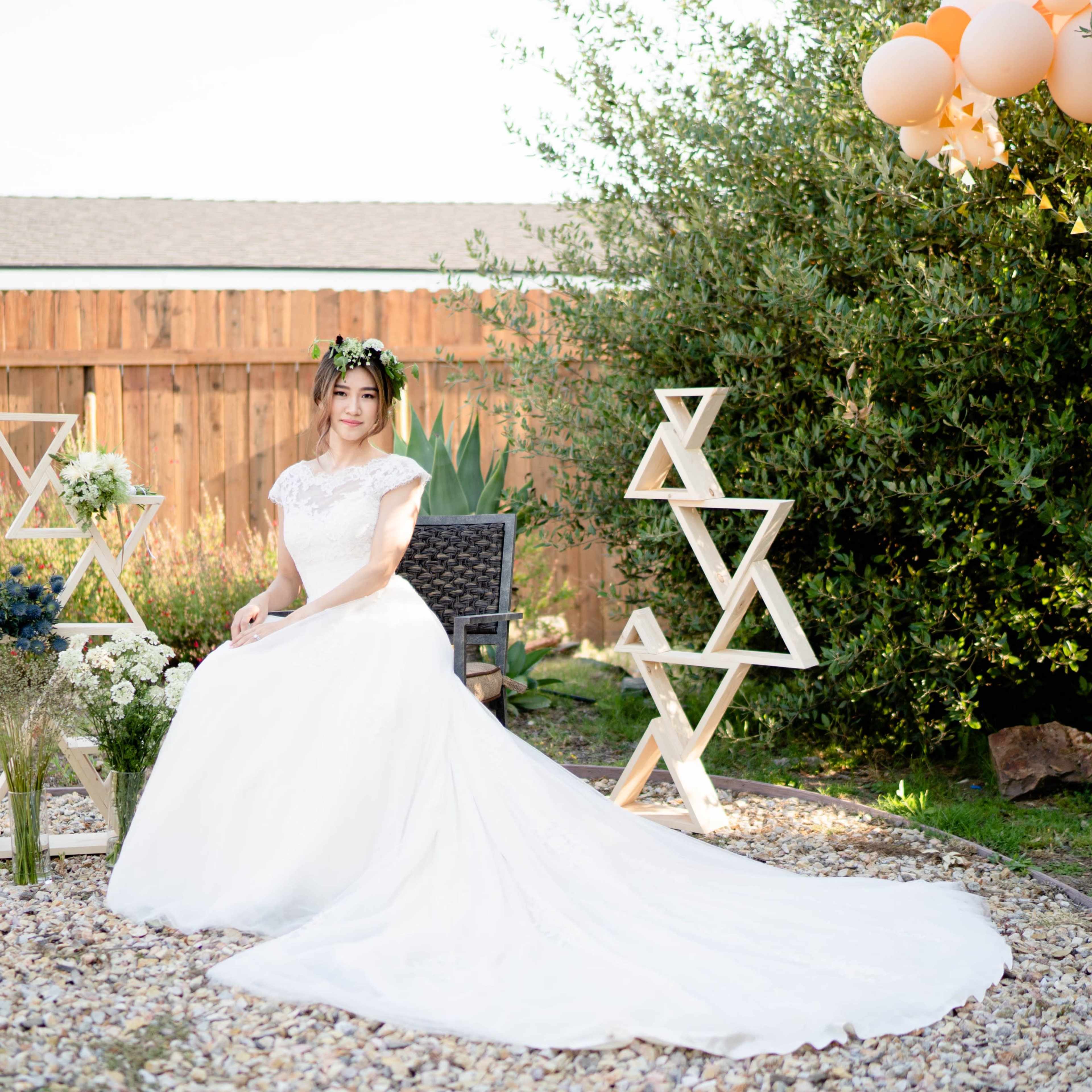 A woman in a white wedding dress sits on a chair in a garden decorated with geometric wooden sculptures and balloons.