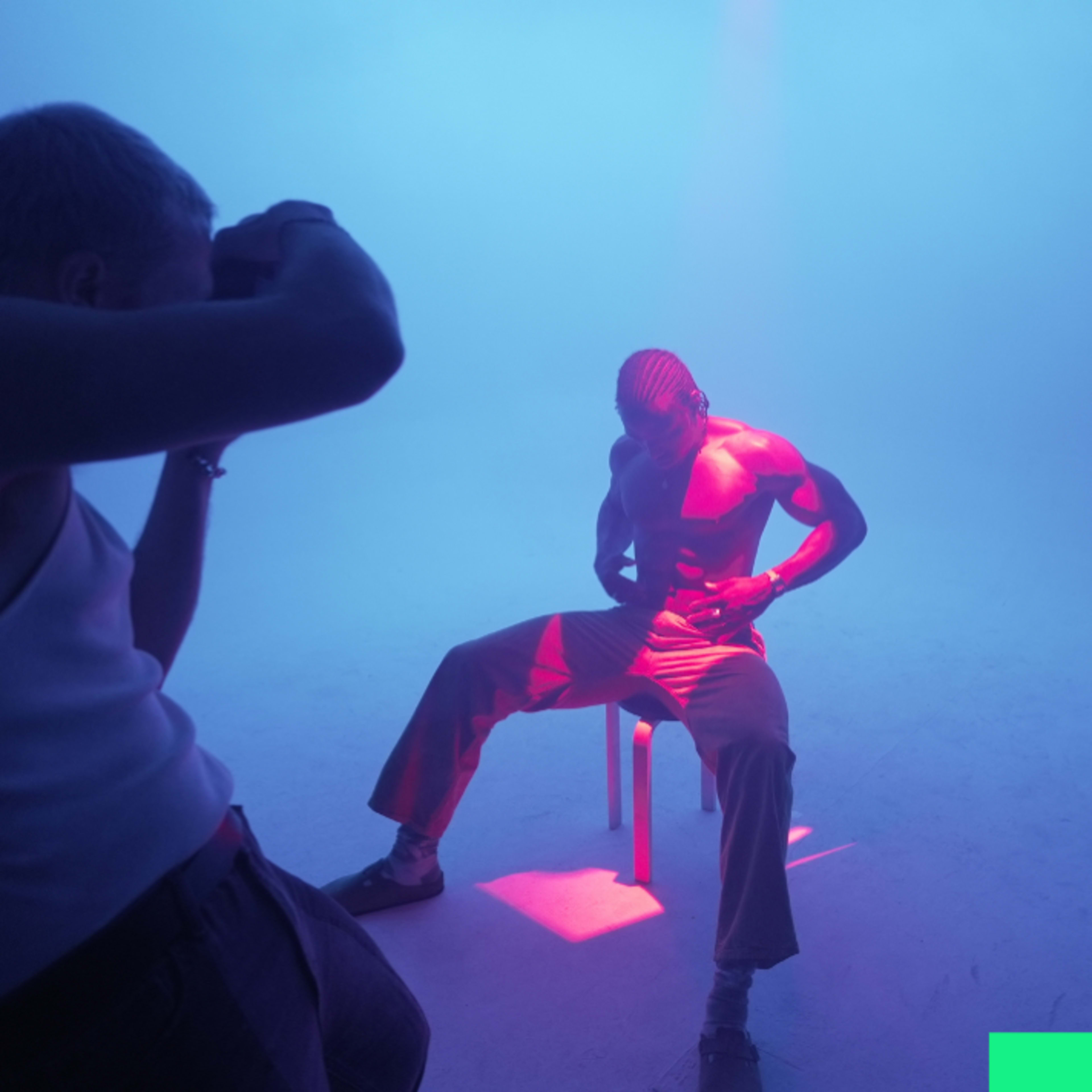 A photographer captures an artistic shot of a male model seated on a stool under colorful lighting in a foggy environment.