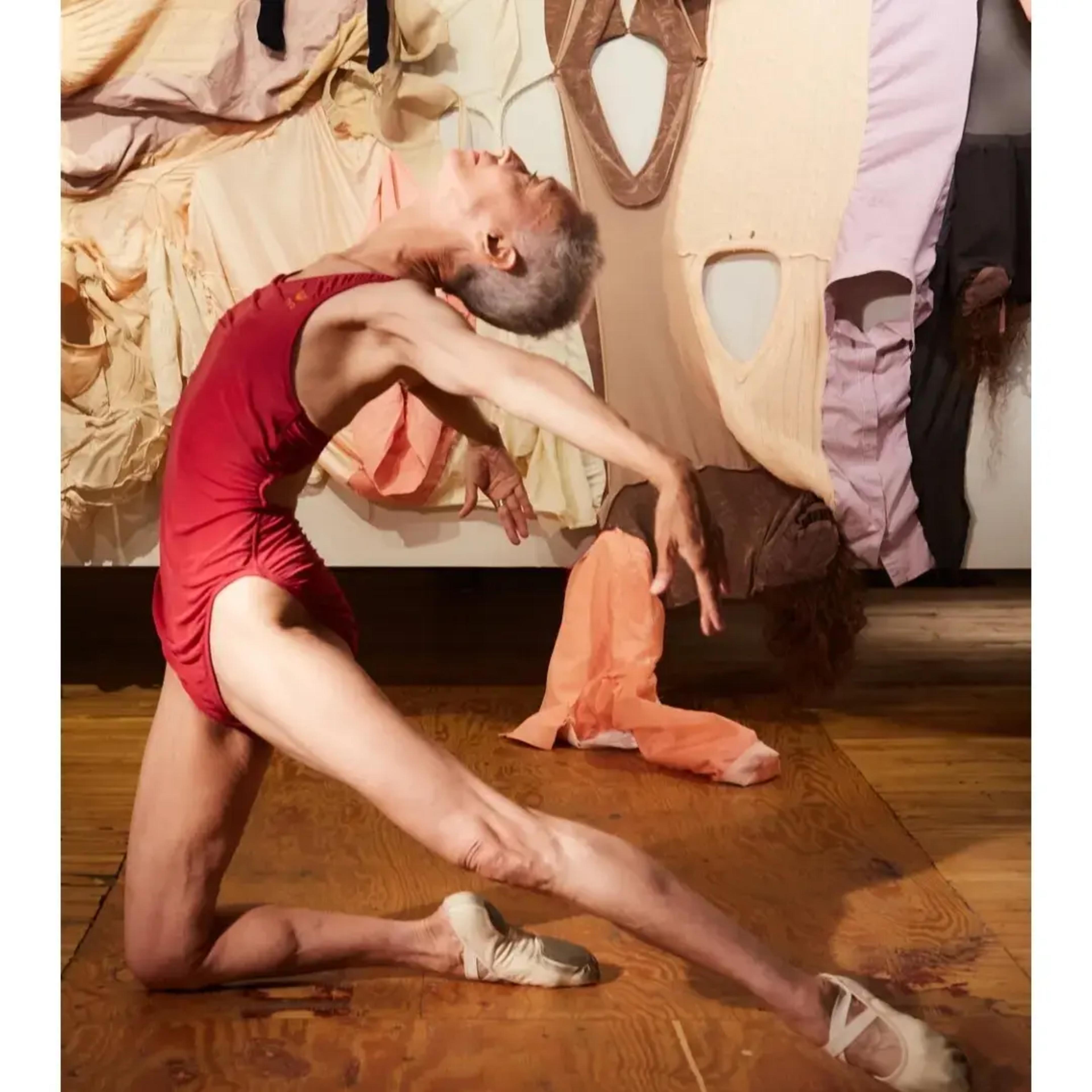 A dancer in a red leotard performs a backbend on a wooden floor in front of a wall adorned with various fabric garments.