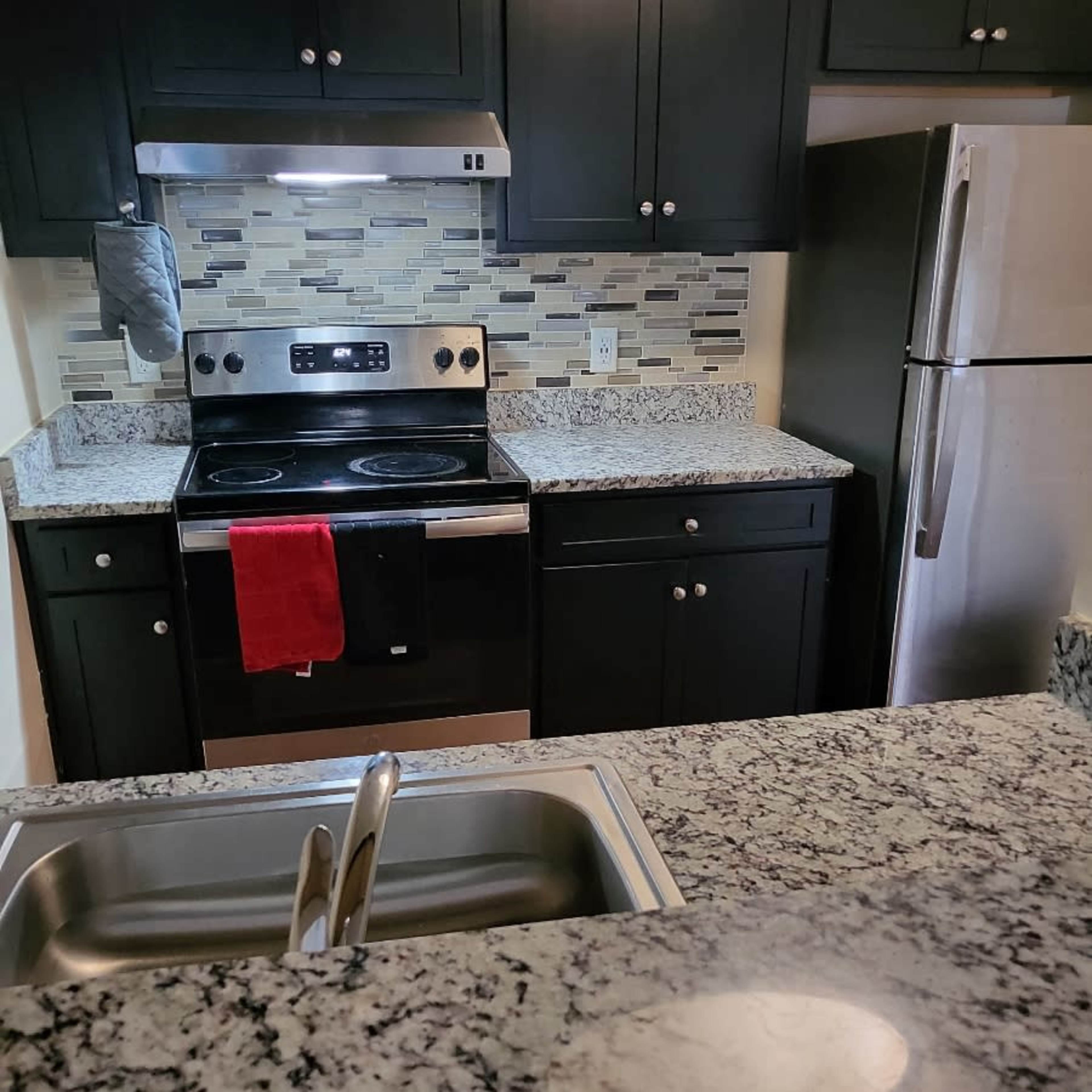 A kitchen with dark cabinets, granite countertops, a stainless steel refrigerator, and an oven with a red dish towel hanging from the handle.