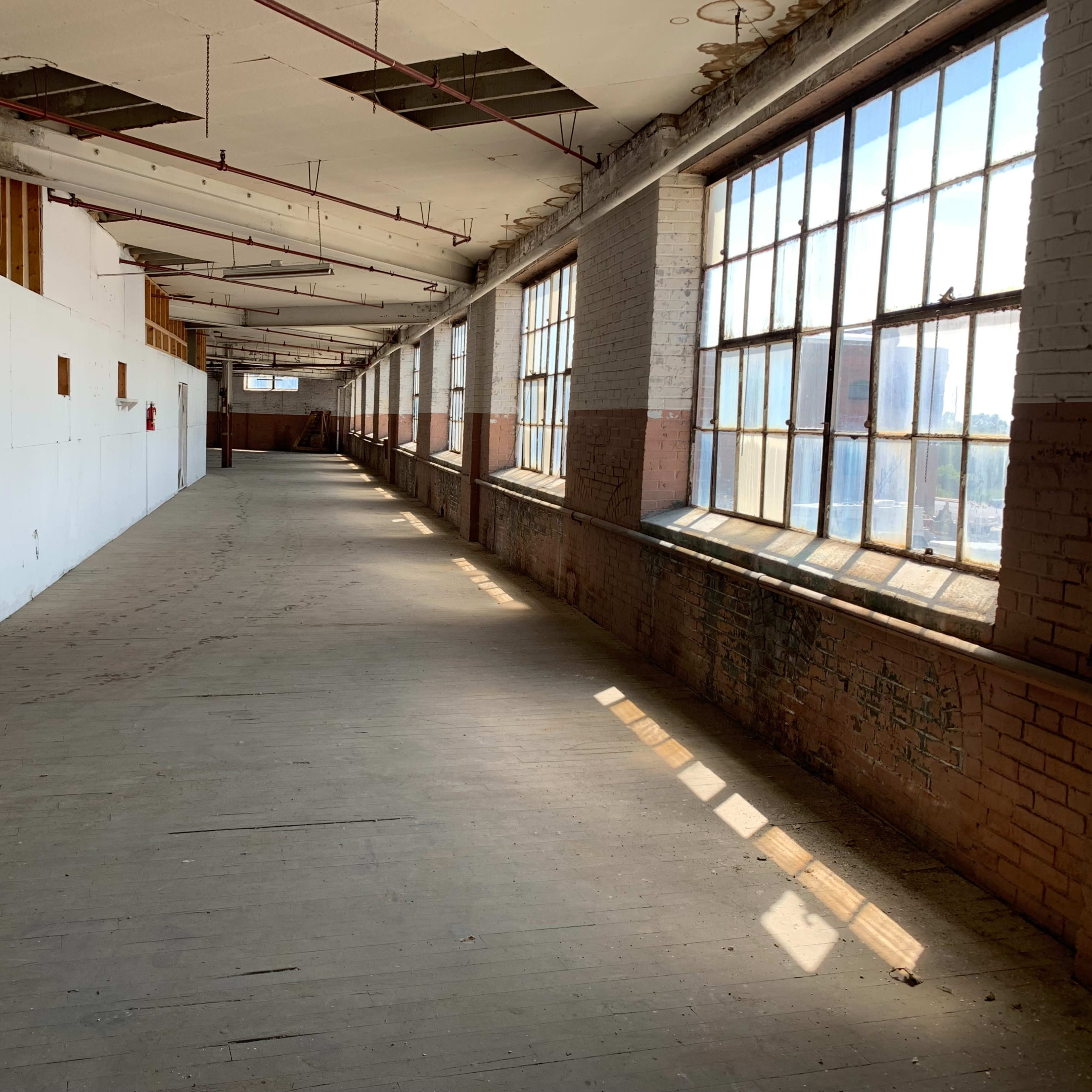 A long, empty corridor with large windows along one side, allowing sunlight to stream in onto the wooden floor.