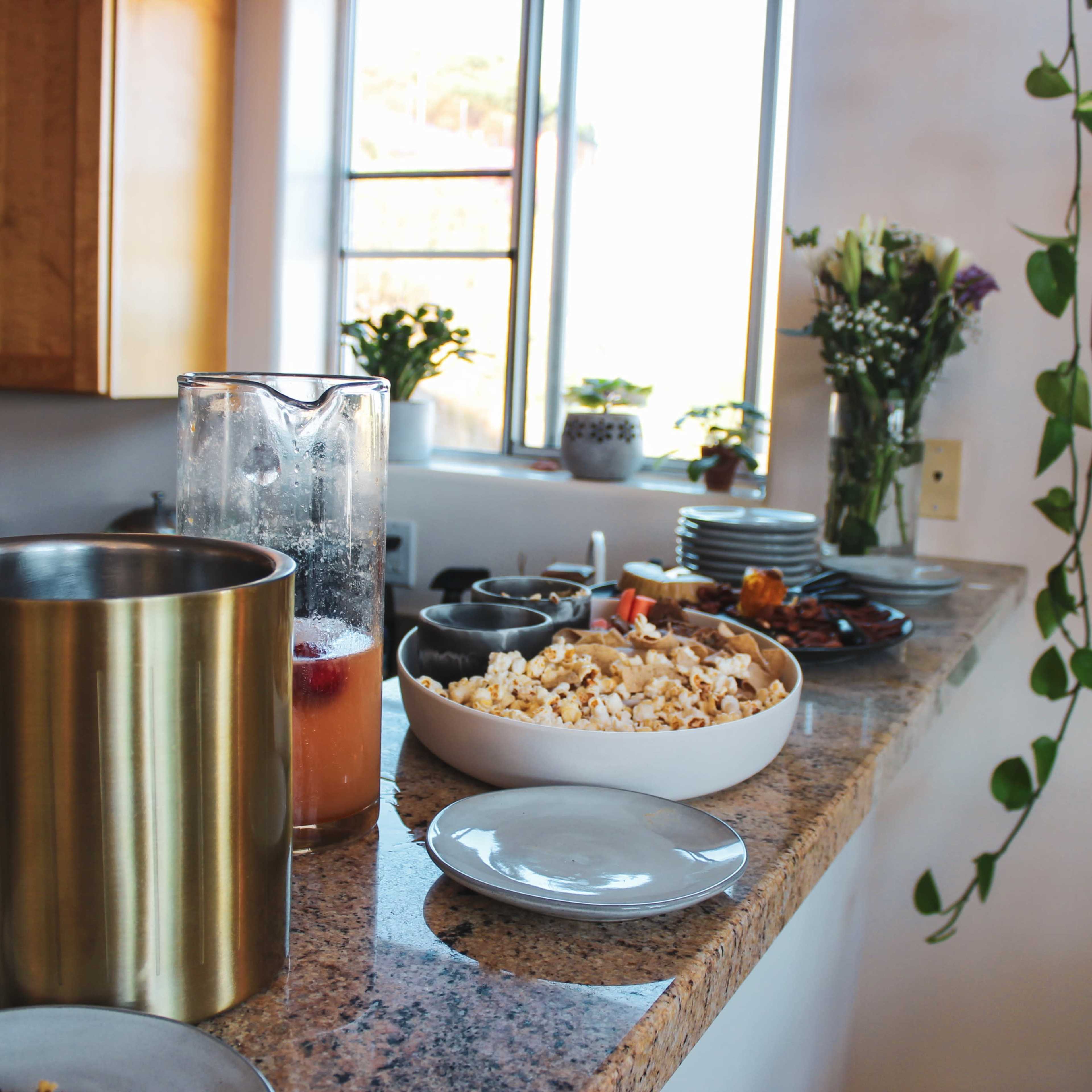A kitchen countertop is filled with a variety of food items, including popcorn, plates, and beverages, alongside a window and plants in the background.