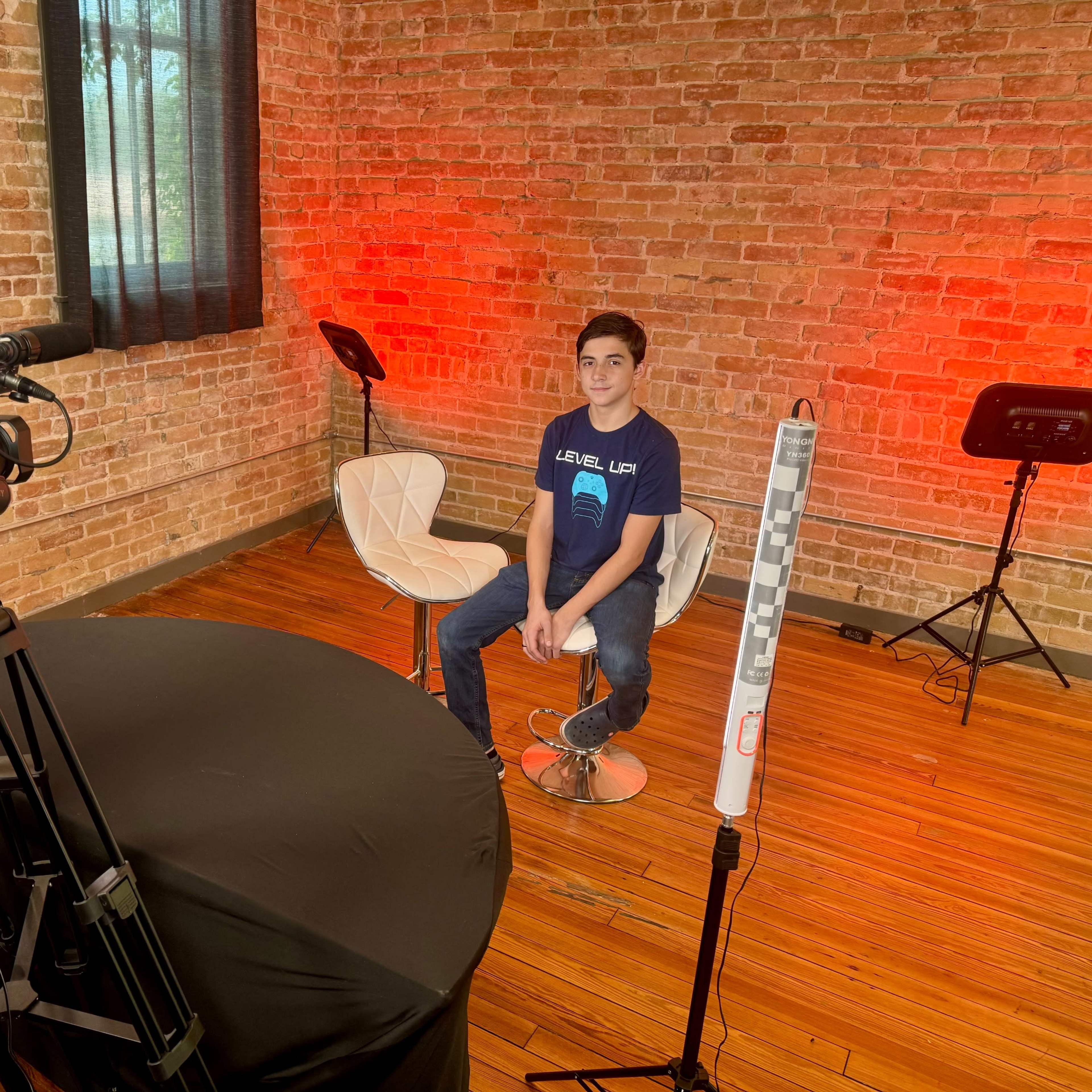 A young boy sits on a white chair in a brick-walled room set up for video recording, with cameras and equipment positioned around him.