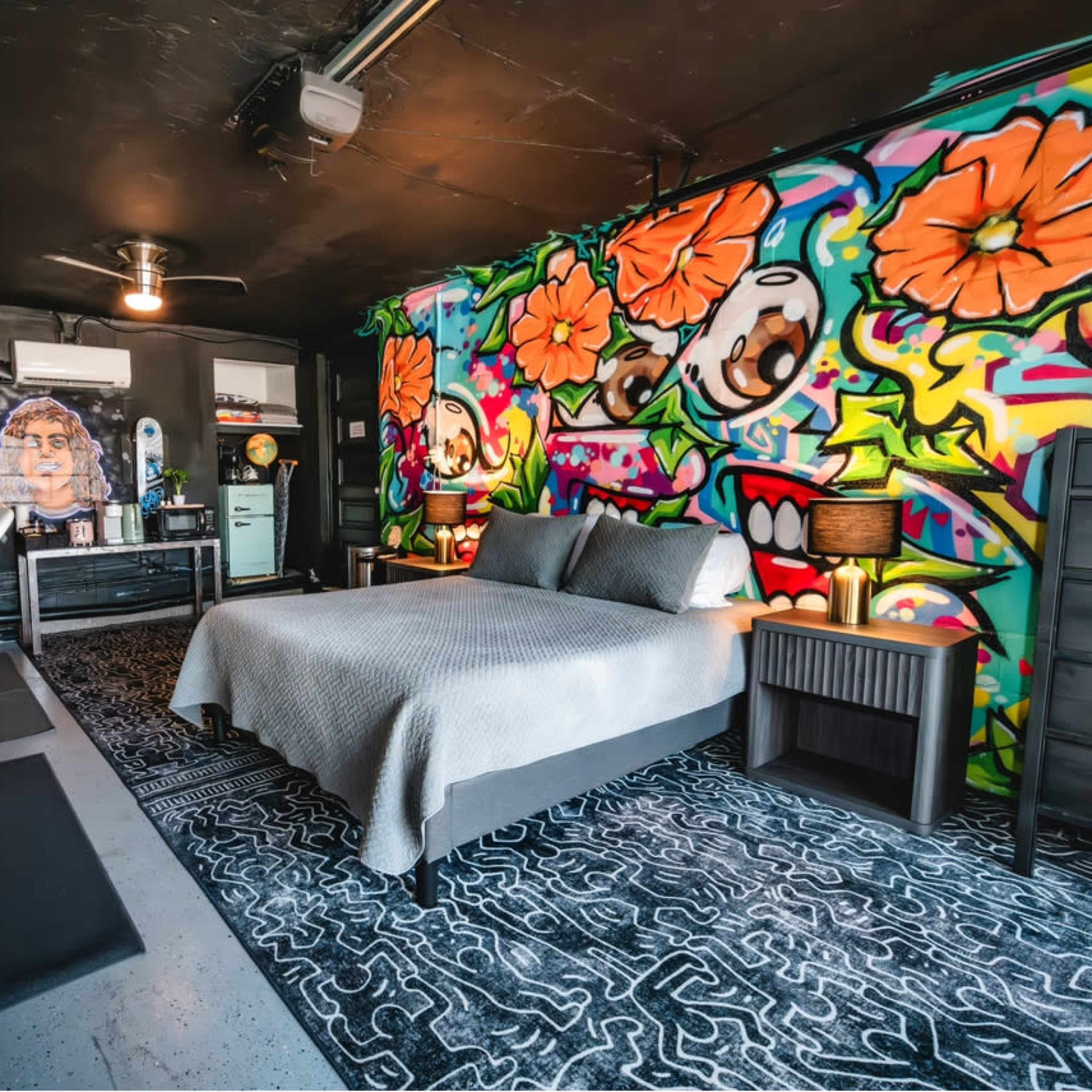 A modern bedroom featuring a bed, colorful mural on the wall, a large rug, a desk area, and a dresser.