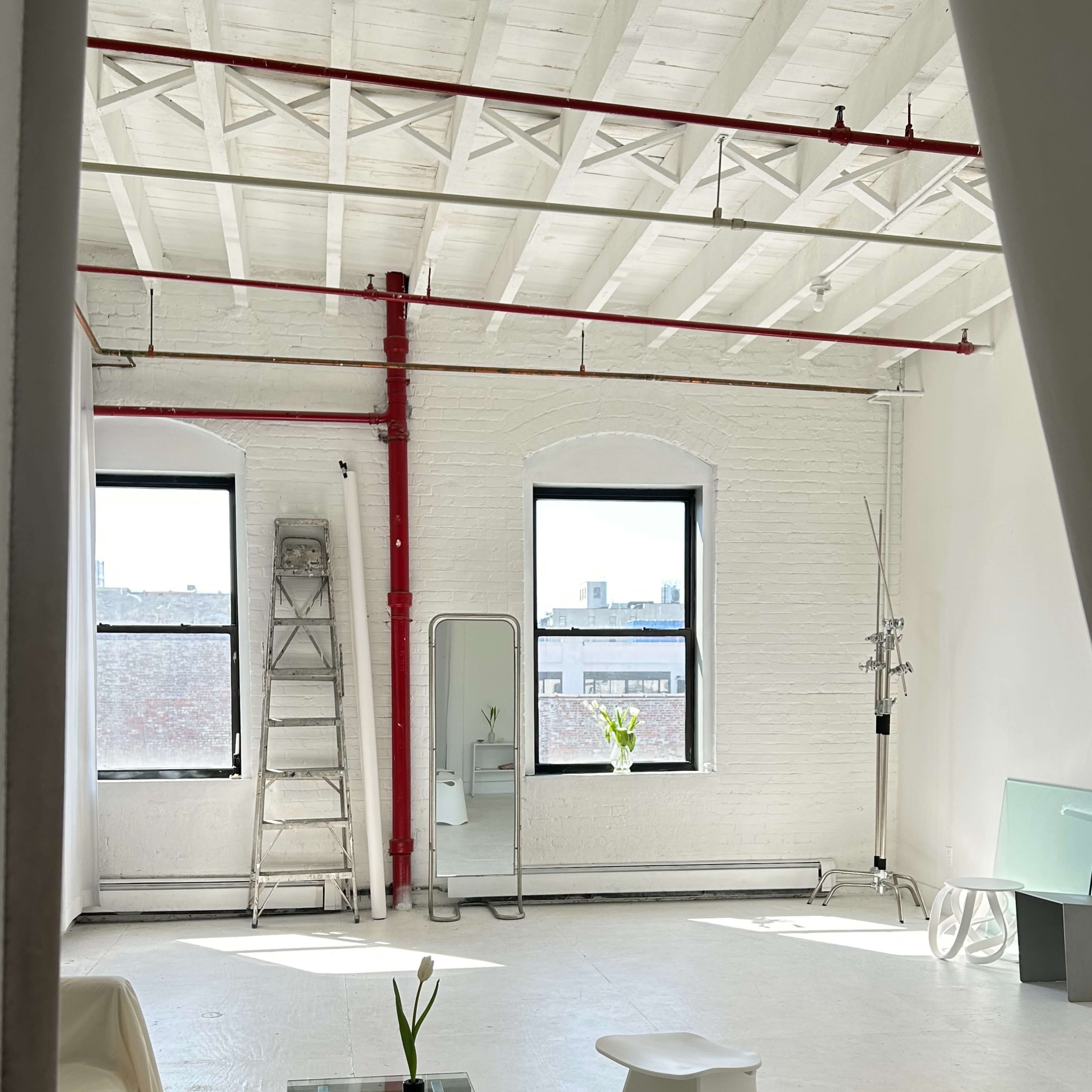 A minimalist interior space with white walls, two windows, a glass table, two white chairs, a ladder, and a potted plant.