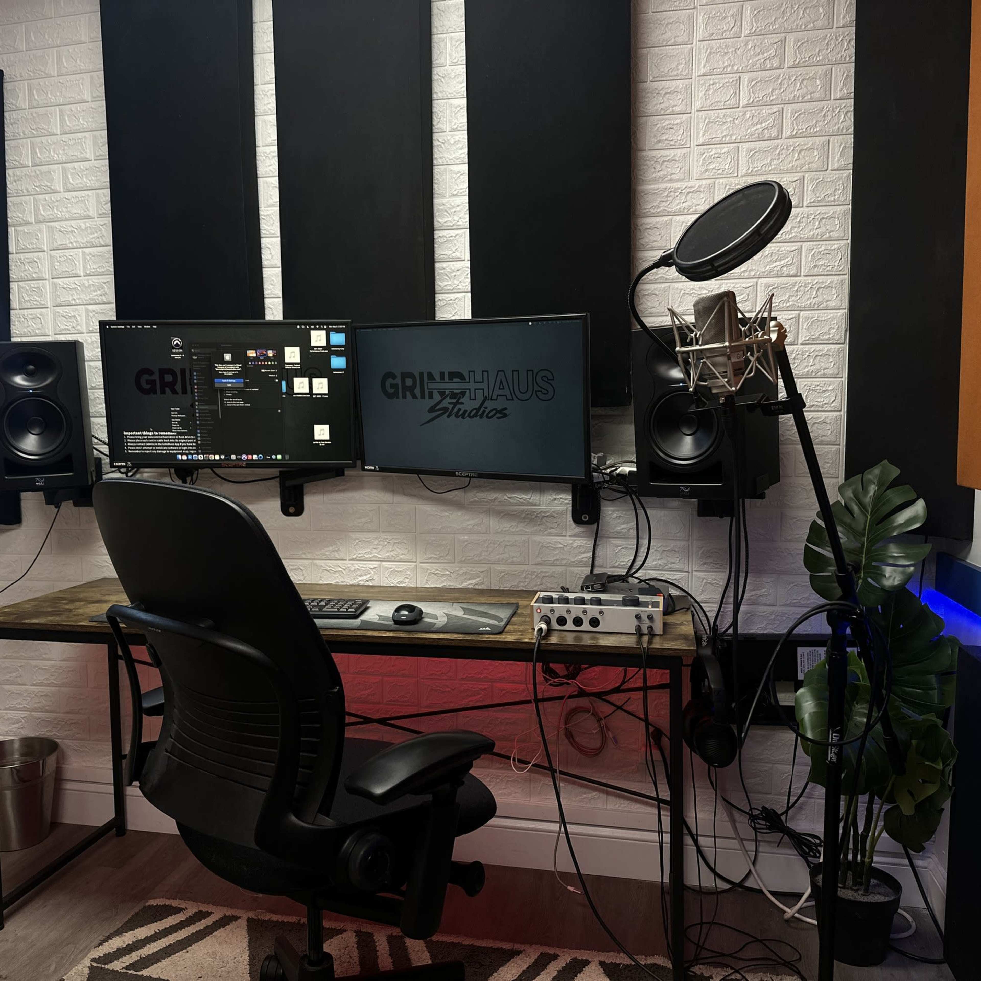 A recording studio with a desk equipped with two monitors, a microphone on a stand, a mixing interface, and soundproof panels on the walls.