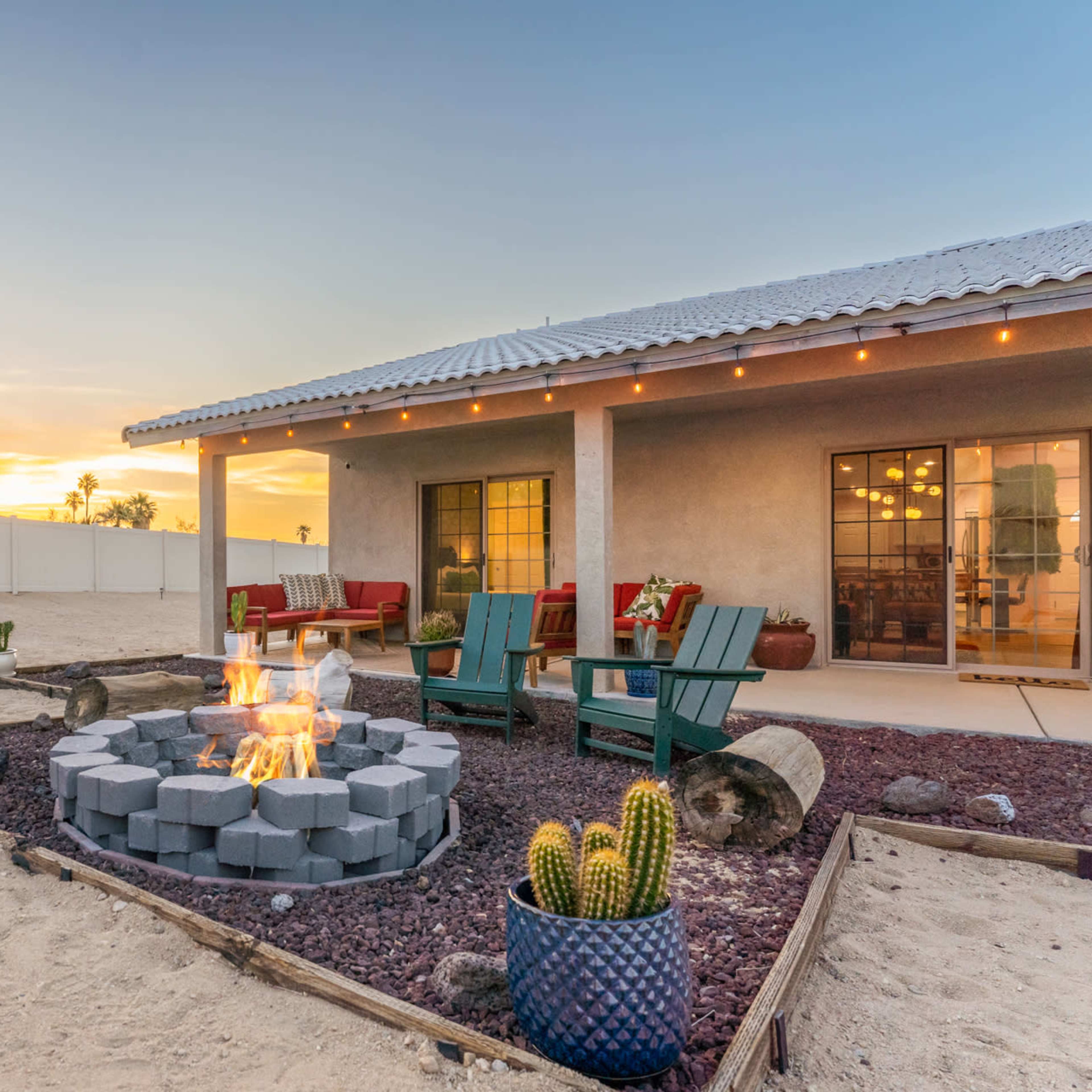 A fire pit surrounded by seating and desert landscaping is illuminated by a sunset in the backyard of a home.