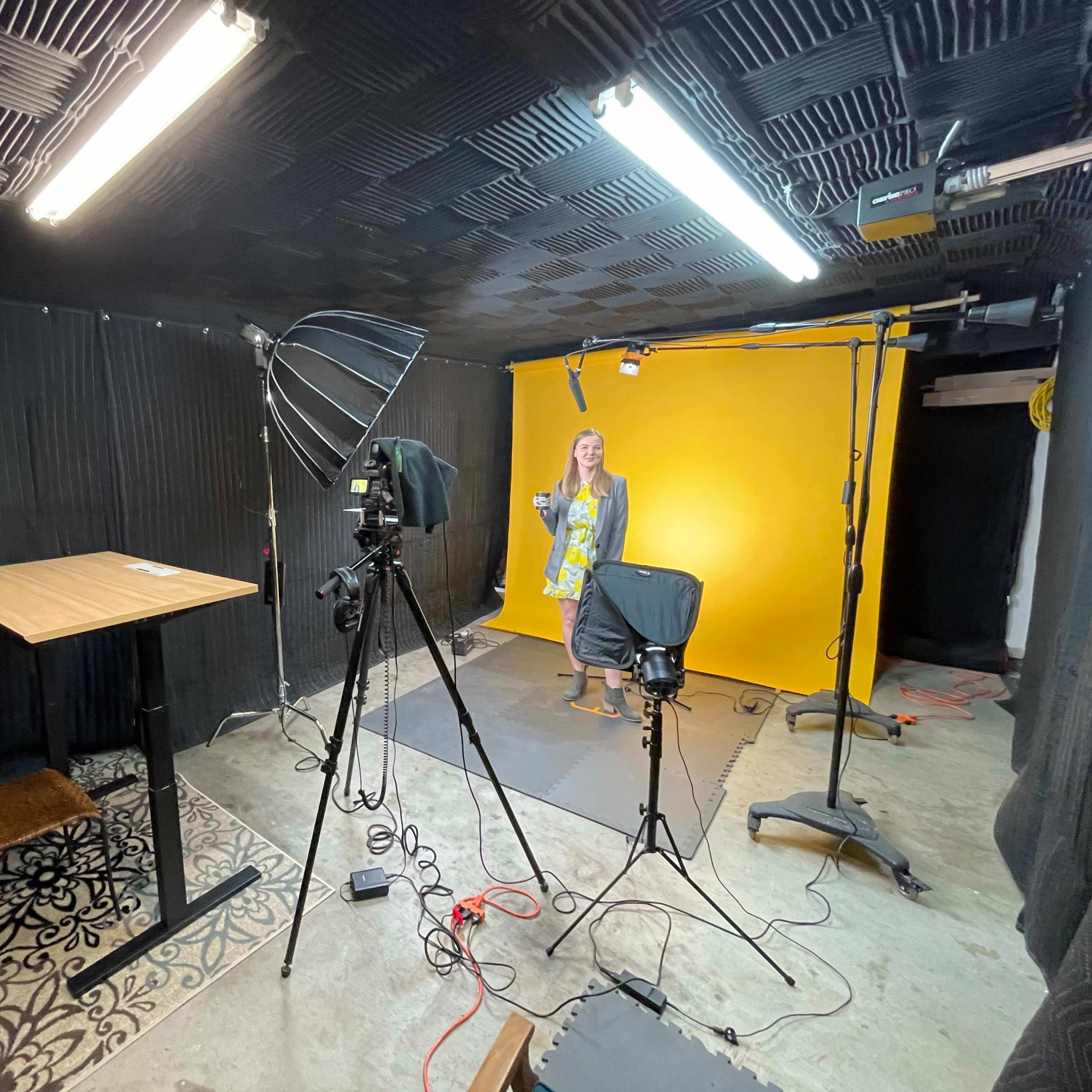 A photography studio is set up with a yellow backdrop, lighting equipment, a camera on a tripod, and a table, while a person stands in the frame.