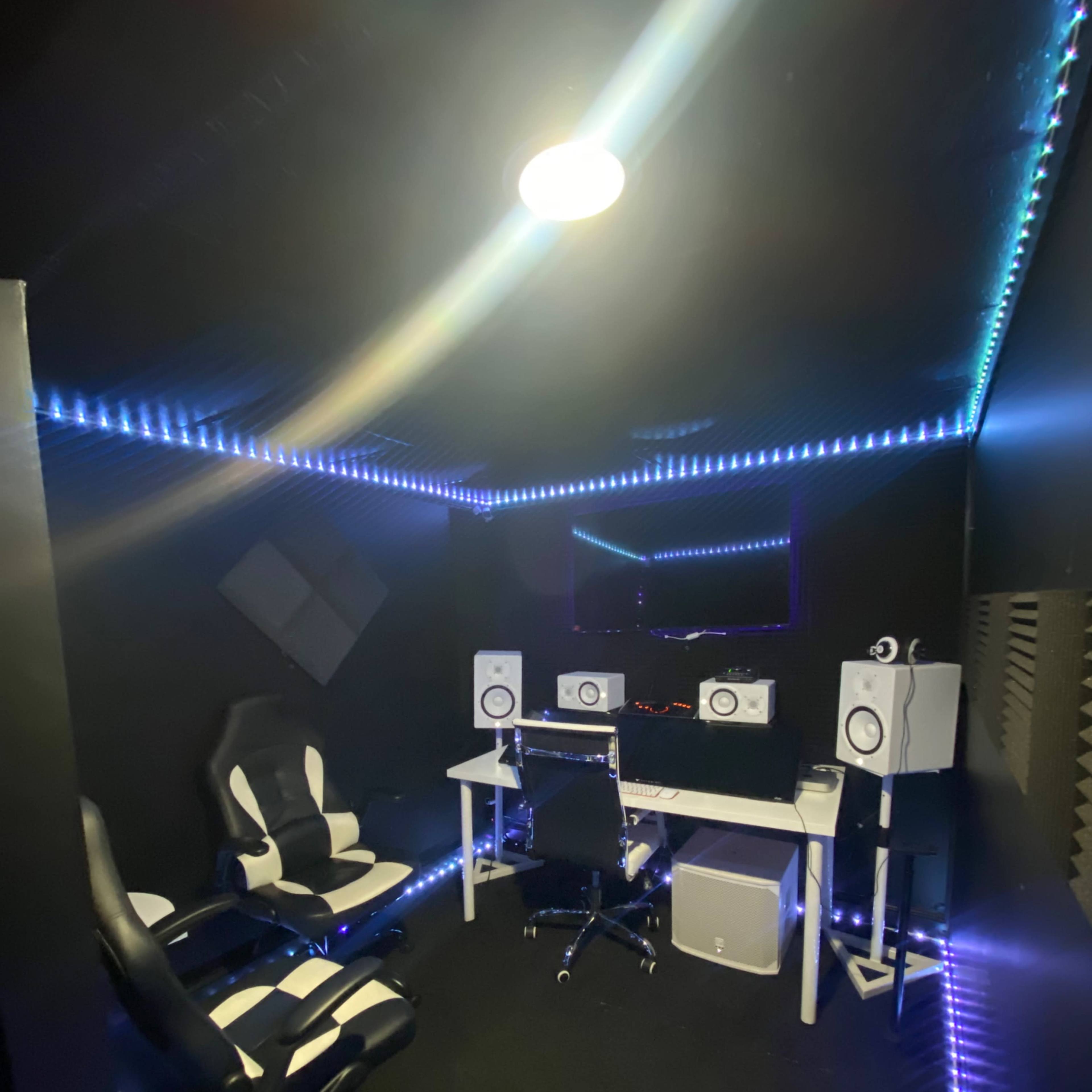 A music recording studio with a black interior, featuring two white monitors, a desk with equipment, and two modern black and white chairs, all illuminated by LED strip lights.